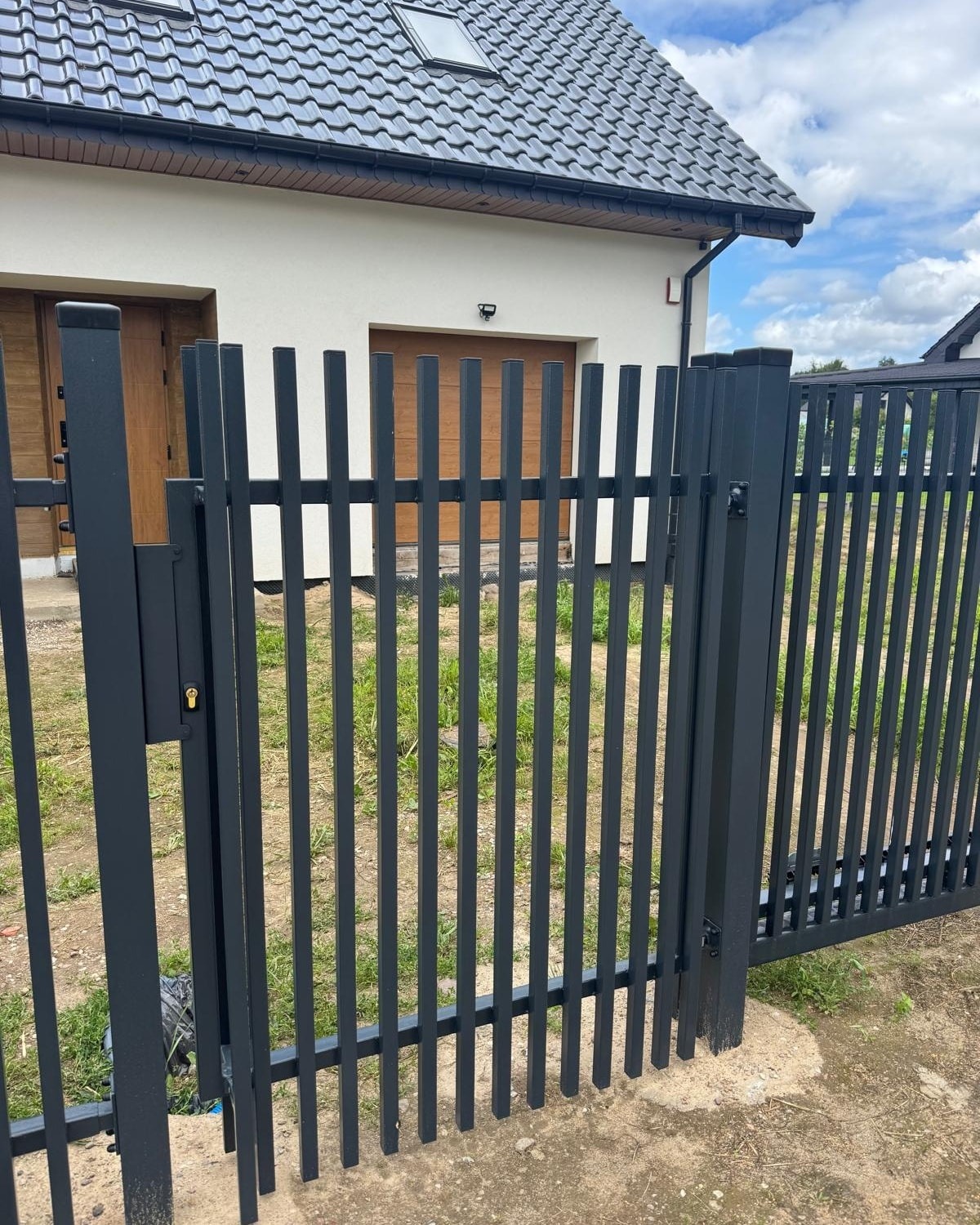 Nowoczesna, grafitowa brama panelowa z furtką, zamontowana przed domem w Płotach. Pionowe profile, minimalistyczny design, widoczny zamek i fragment budynku w tle.