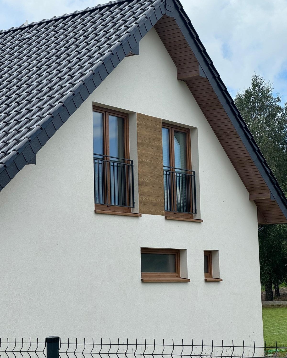 Biały dom z ciemną dachówką i drewnianymi oknami. Czarne, metalowe balustrady balkonowe przy oknach na piętrze. U dołu widoczne ogrodzenie z siatki.