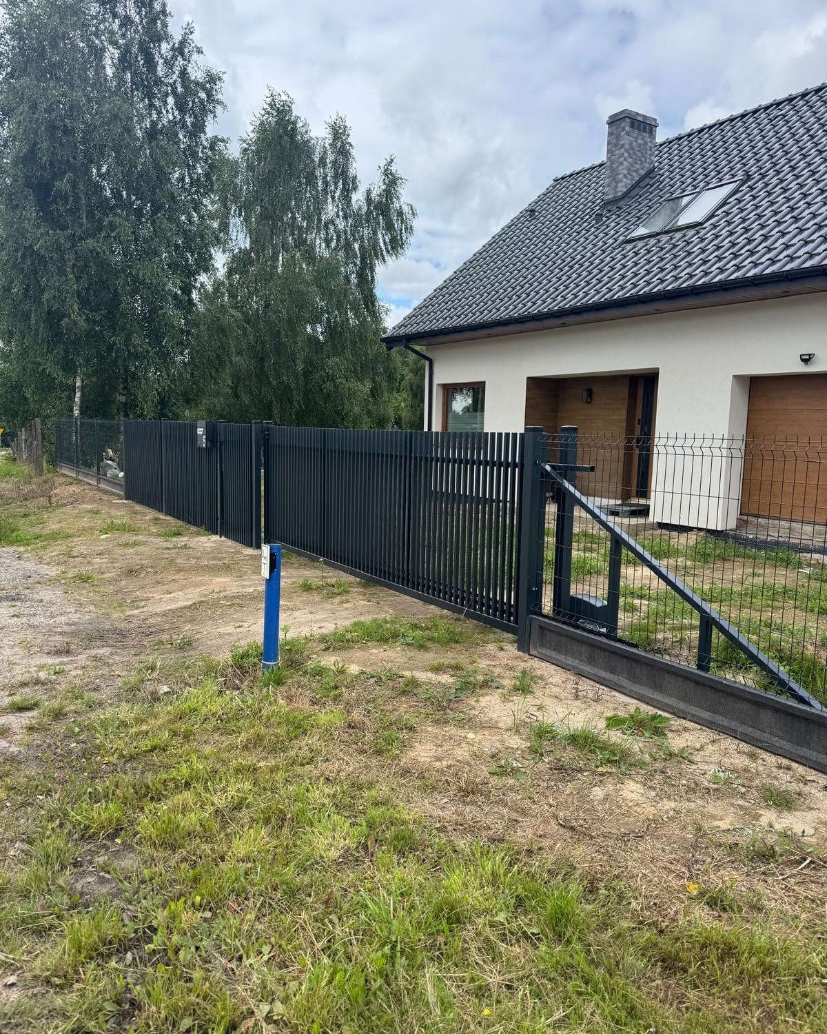 Nowoczesne, grafitowe ogrodzenie metalowe z bramą wjazdową, widoczne na tle domu z szarą dachówką i zielenią. Słupki ogrodzeniowe z niebieskim akcentem.