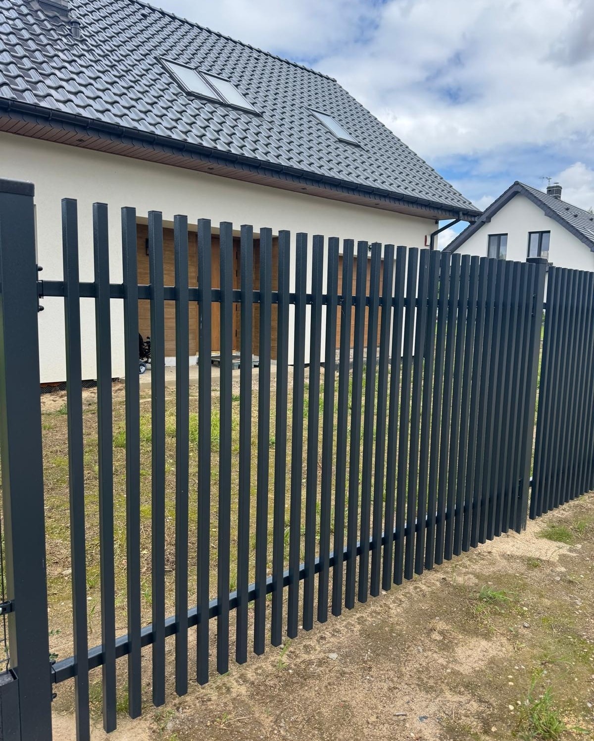 Nowoczesne, pionowe ogrodzenie metalowe w kolorze grafitowym, z widocznymi dwoma domami w tle, w słoneczny dzień. Minimalistyczny design.
