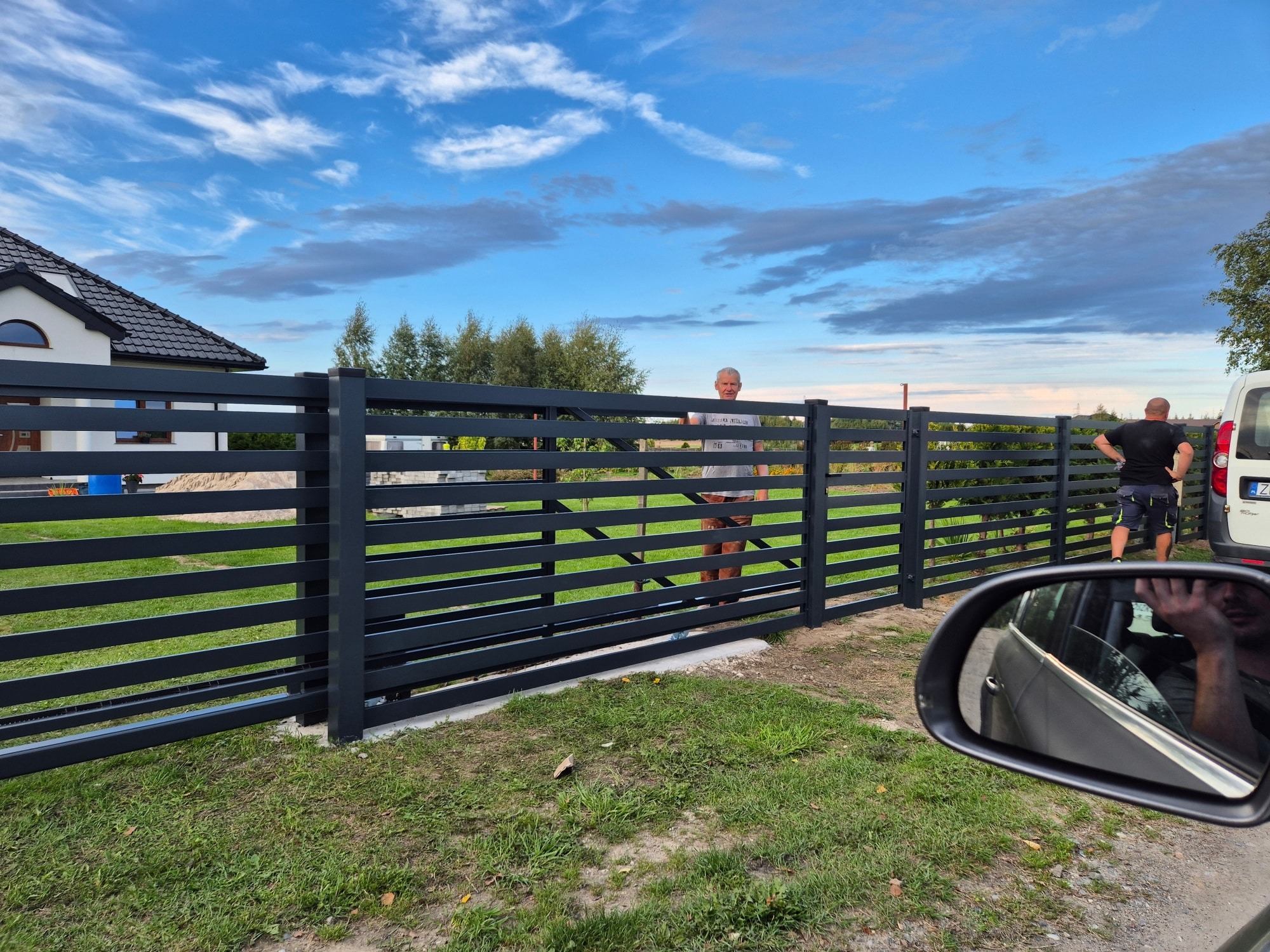 Nowoczesne, poziome ogrodzenie metalowe w kolorze antracytowym, z widocznymi dwoma mężczyznami w tle, na tle domu i błękitnego nieba z chmurami.