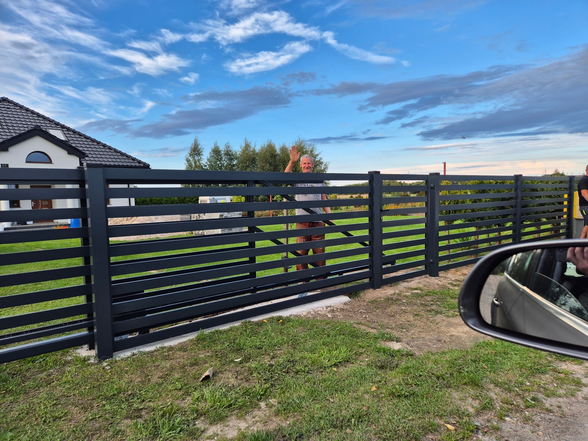 Nowoczesne, poziome ogrodzenie panelowe w kolorze grafitowym, z widoczną bramą i uśmiechniętym mężczyzną machającym ręką, na tle domu i błękitnego nieba.