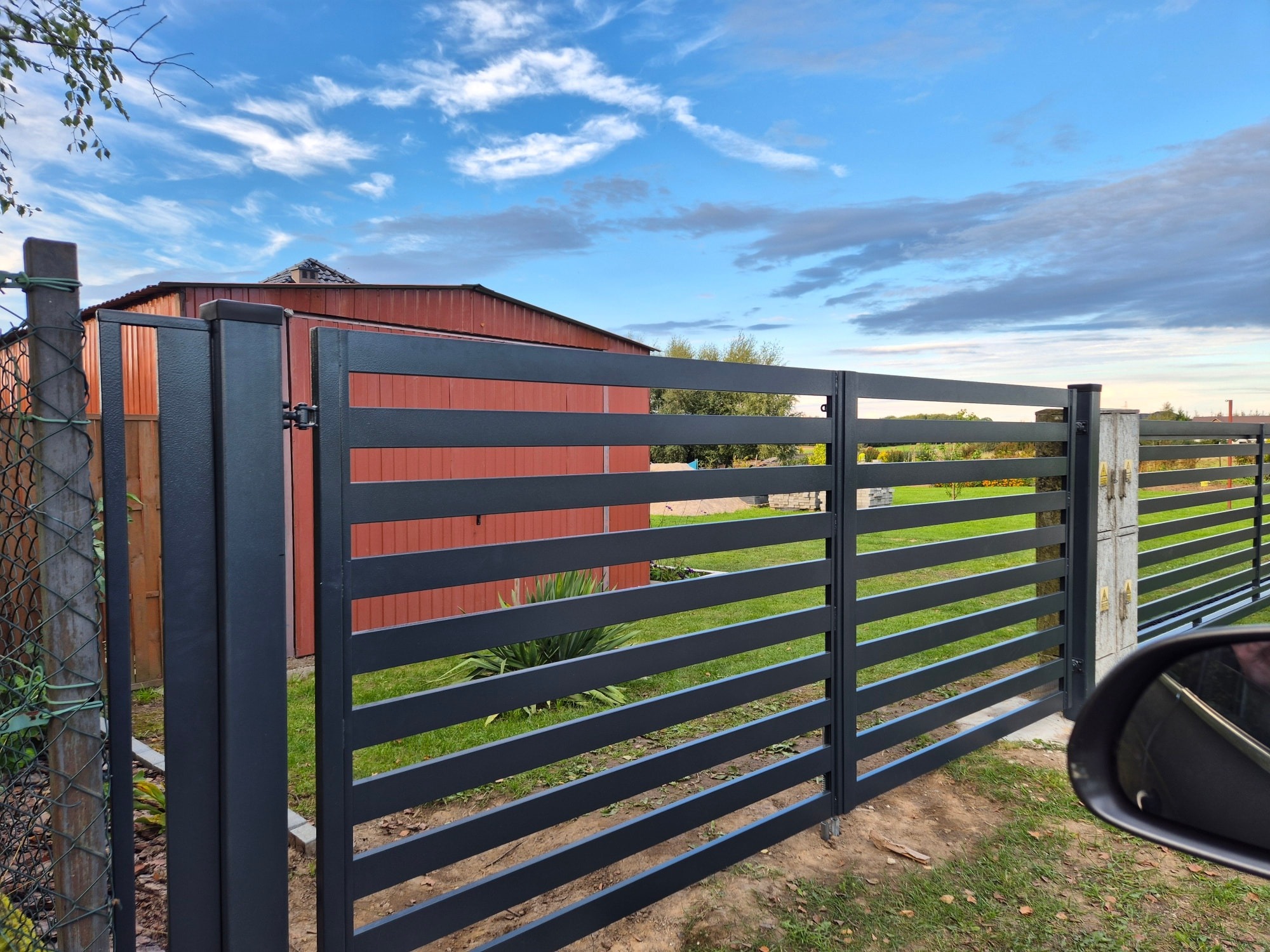 Nowoczesne, poziome ogrodzenie panelowe w kolorze grafitowym, z betonowymi słupkami, na tle czerwonego budynku i błękitnego nieba z chmurami. Widok zza lusterka samochodu.