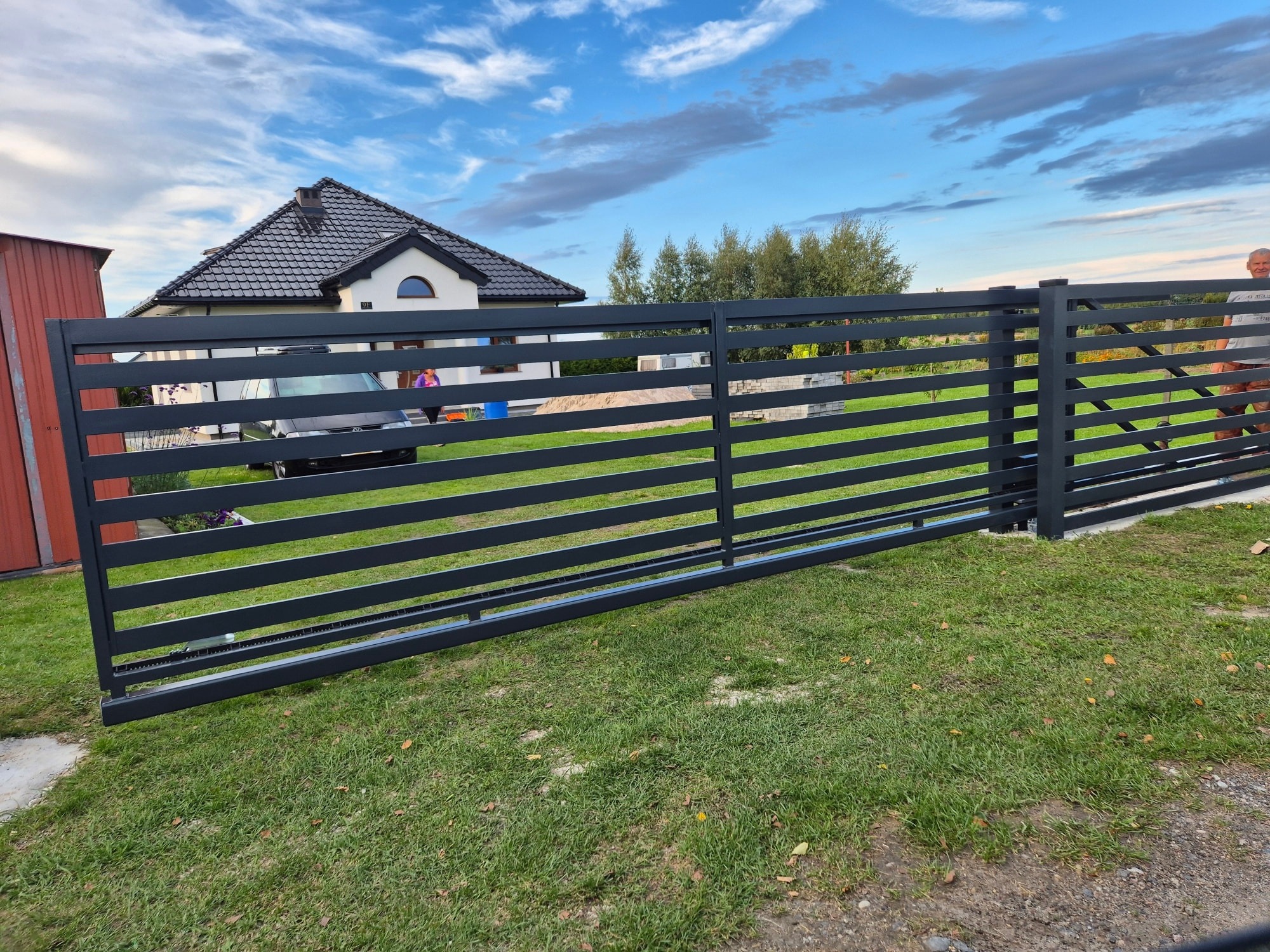 Nowoczesne, poziome ogrodzenie posesji z szarych, metalowych paneli na tle domu z ciemnym dachem i błękitnego nieba z chmurami. Widoczny mechanizm bramy przesuwnej.