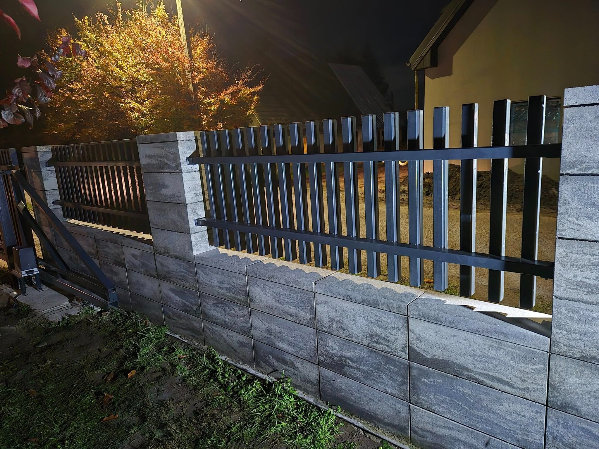 Nowoczesne ogrodzenie z szarej kostki i metalowych paneli w kolorze grafitowym, oświetlone nocą, z widocznym automatem do bramy przesuwnej.