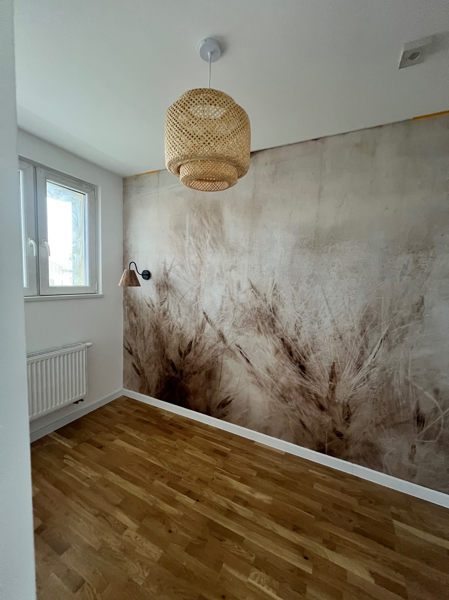Pokój po remoncie: drewniana podłoga, ściana z motywem roślinnym, pleciona lampa wisząca i kinkiet. Nowoczesne wykończenie wnętrza z naturalnymi akcentami.
