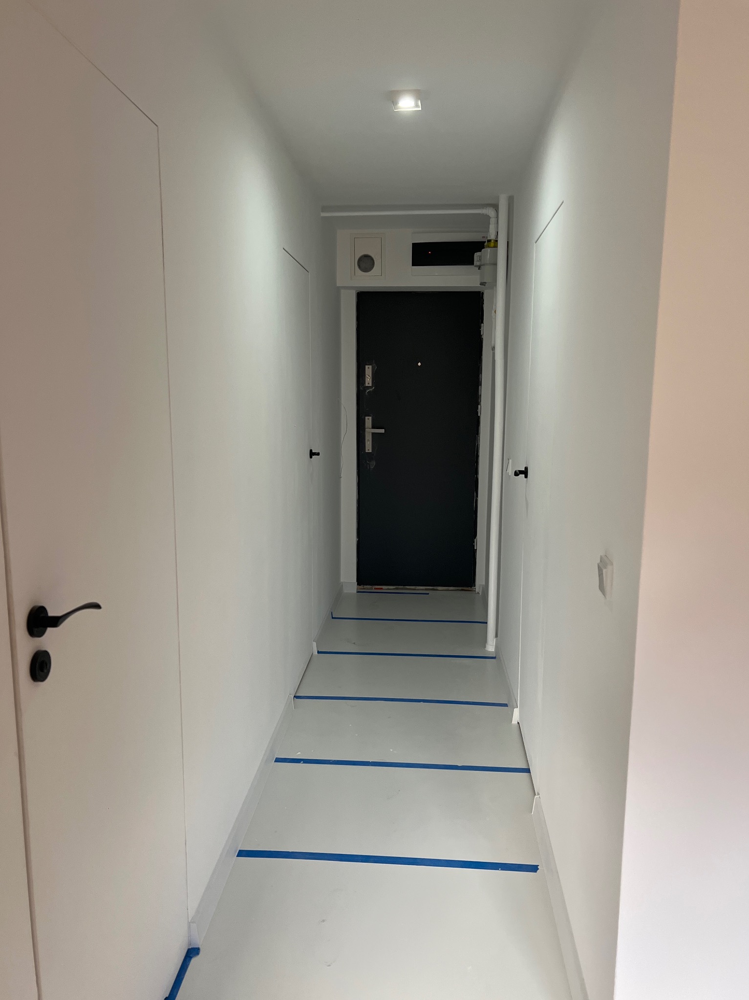 Korytarz z ukrytymi drzwiami w kolorze białym, zabezpieczona podłoga taśmą malarską. Minimalistyczny design, czarne klamki, oświetlenie sufitowe.