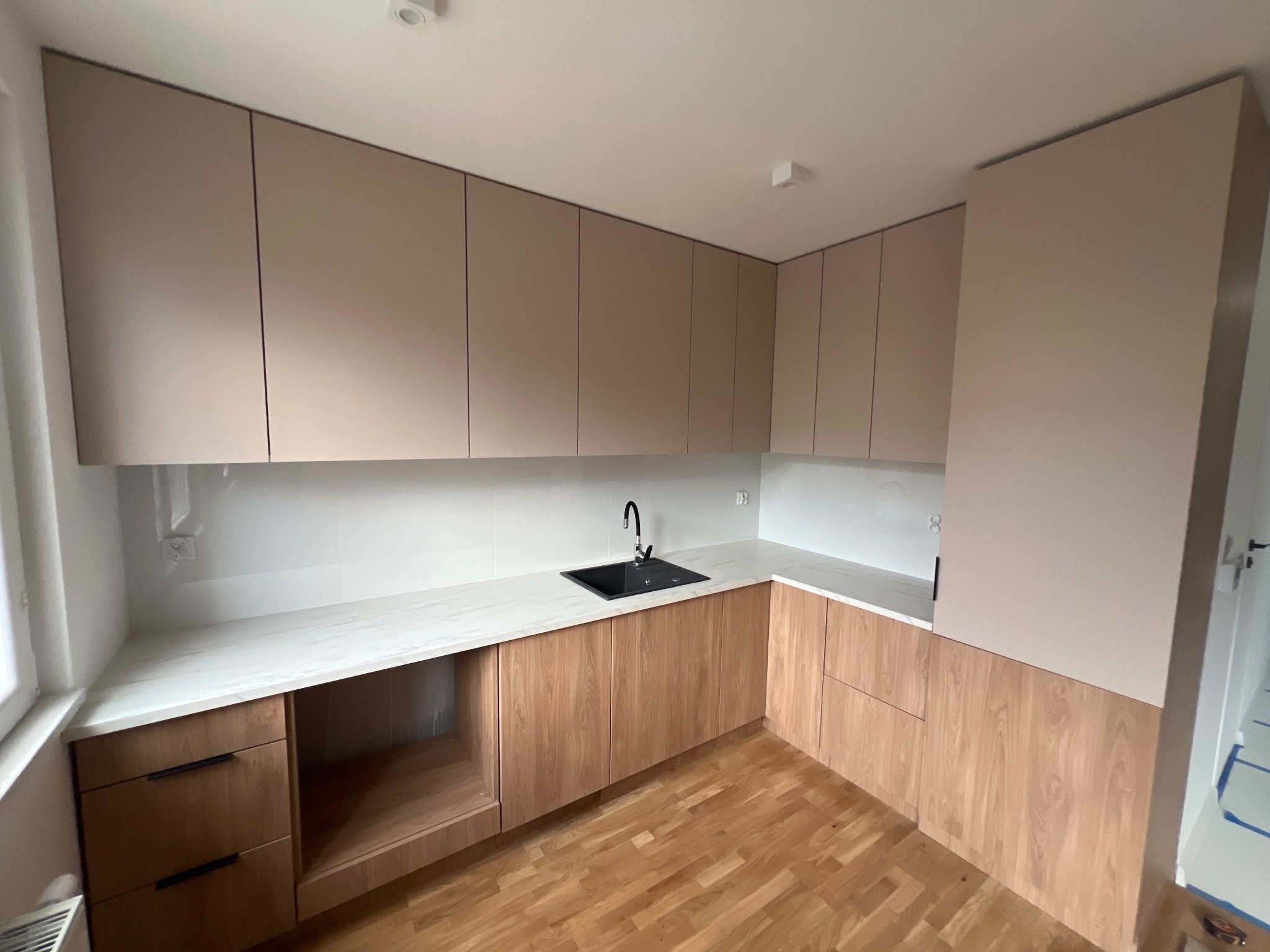 Nowoczesna kuchnia z drewnianymi szafkami dolnymi i beżowymi górnymi, biały blat i ściana, czarny zlew, parkiet. Widok z perspektywy, ukończony montaż.