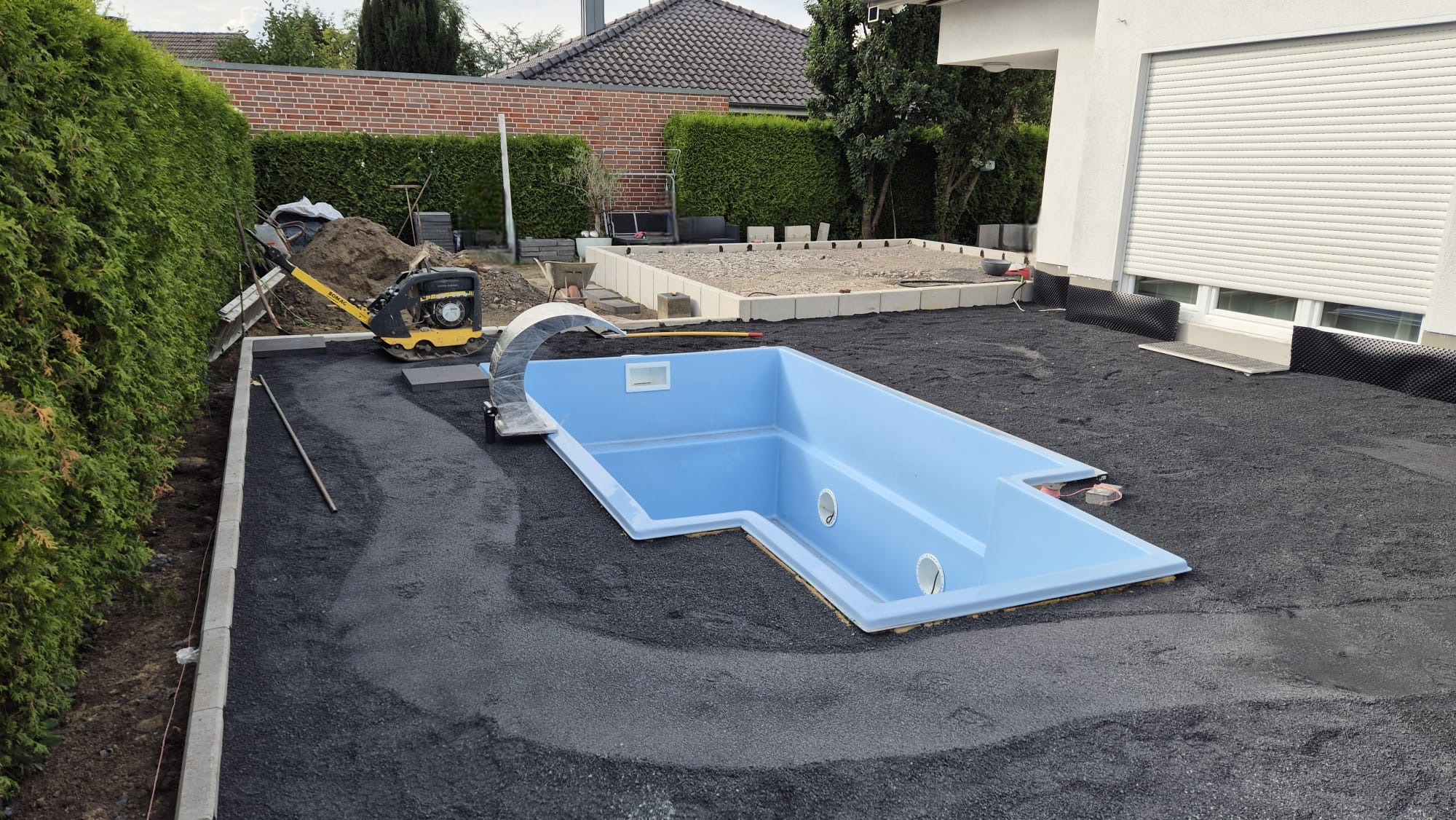 Niebieski basen w ziemi, przygotowany teren z zagęszczarką, obrzeża z kostki, widok na dom i zielony żywopłot. Etap budowy basenu.