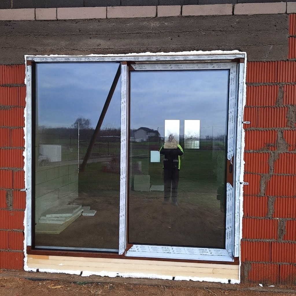 Montaż okna dwuszybowego w surowym murze z czerwonej cegły. Widoczne pianowanie i odbicie osoby w szybie. Okno z brązową ramą wewnętrzną.