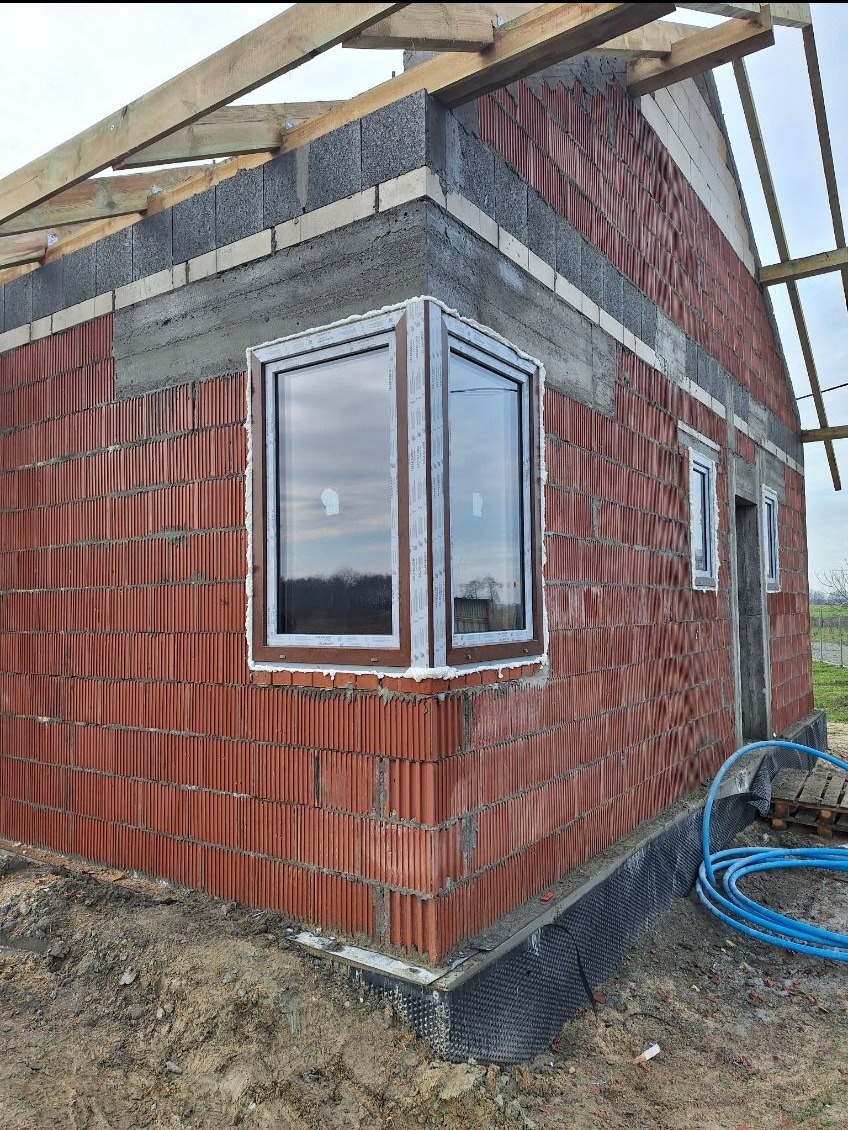 Montaż okna narożnego w surowym budynku z czerwonej cegły. Widoczne drewniane elementy konstrukcji dachu i niebieski wąż ogrodowy na ziemi.