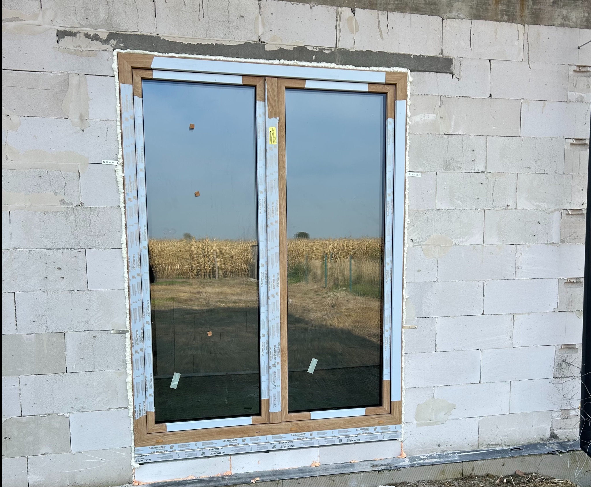 Montaż okna z drewnopodobną ramą w surowym murze. Widoczne pianowanie i listwy montażowe. W szybie odbija się pole kukurydzy.