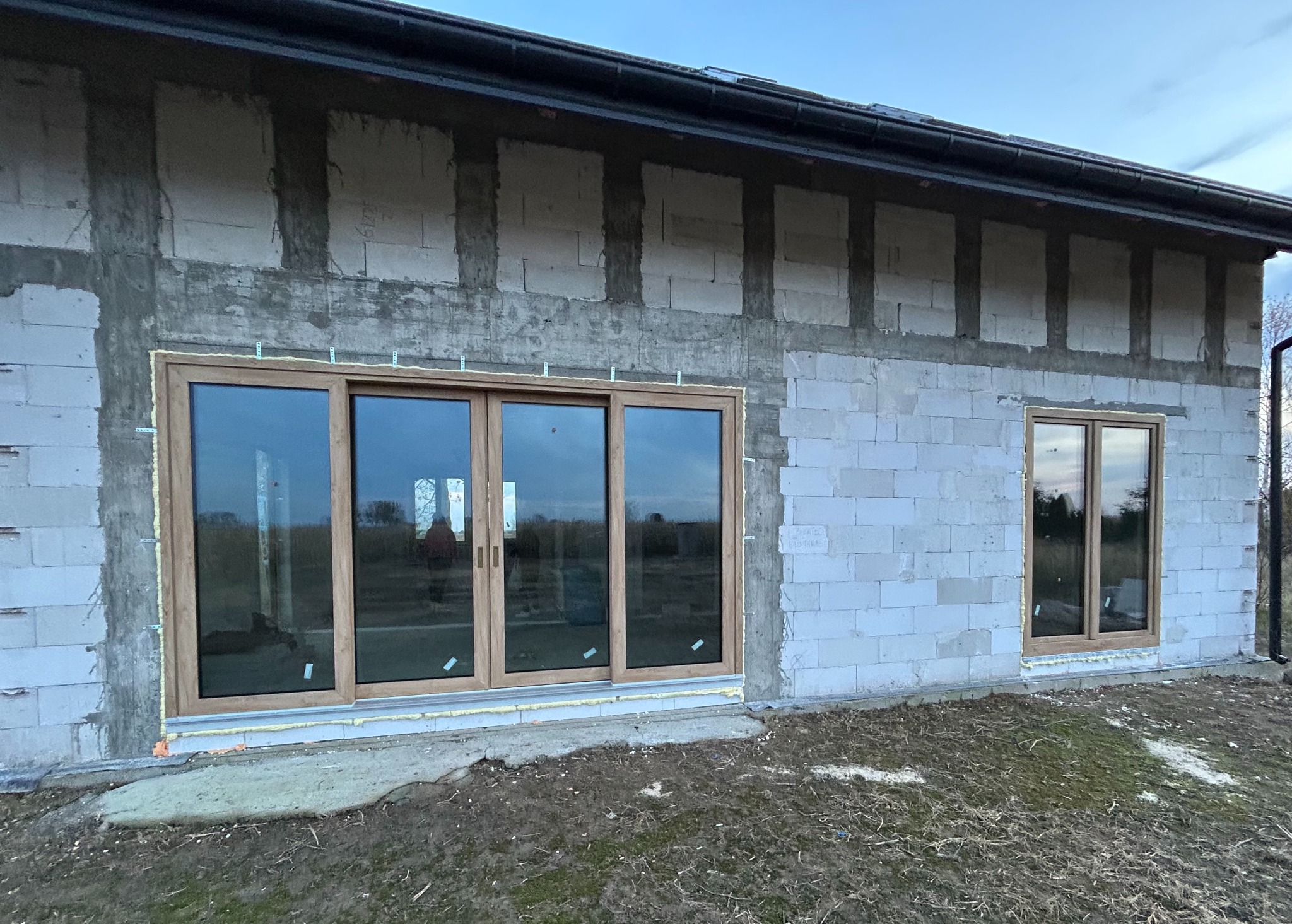 Montaż okien przesuwnych i standardowego okna w surowym budynku z betonu komórkowego. Widoczne pianowanie i mocowania. Na szybach odbija się sylwetka osoby.