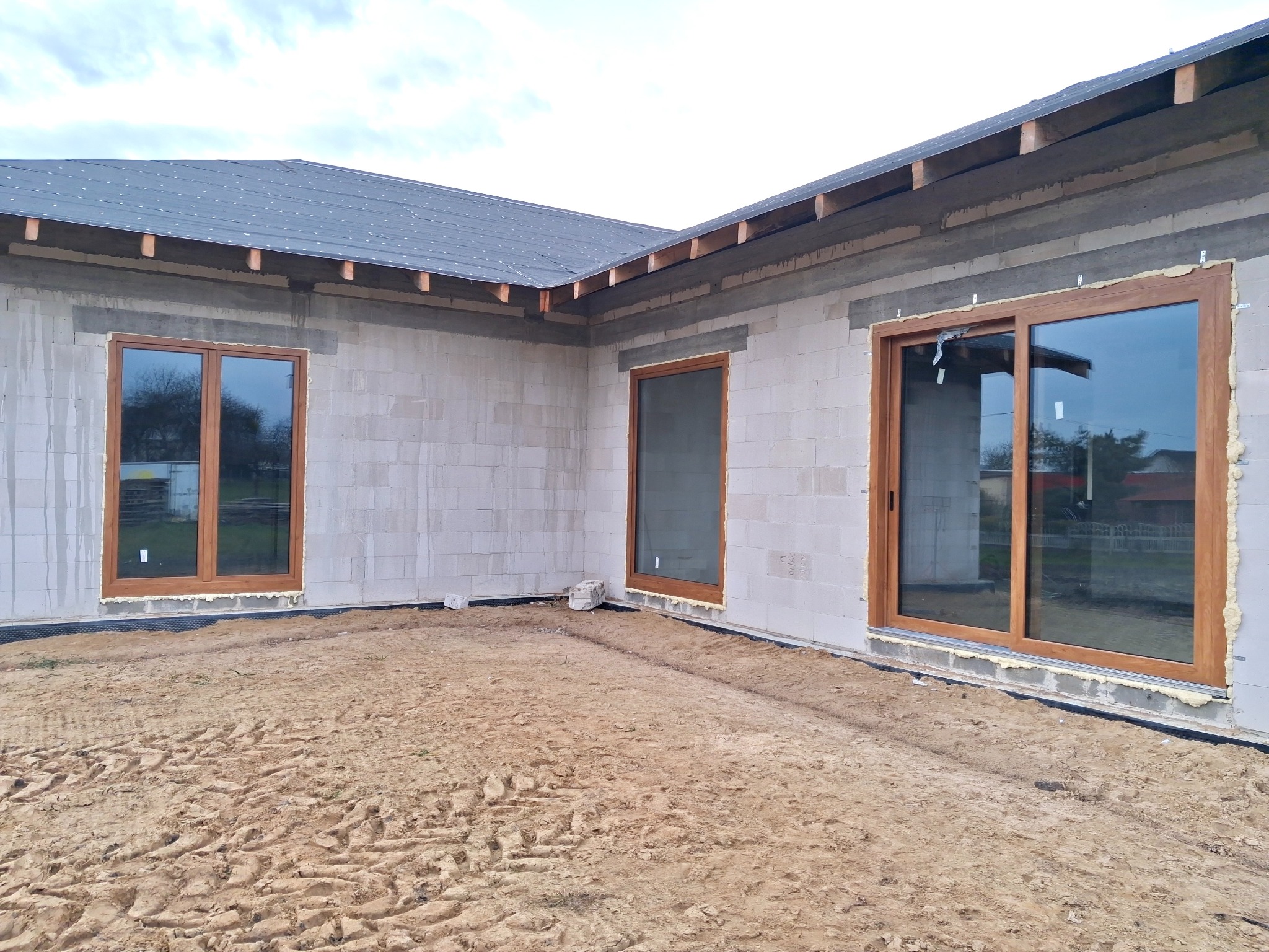 Montaż okien PCV w surowym budynku. Widoczne trzy okna z brązowymi ramami, osadzone w ścianach z betonu komórkowego. Teren wokół budynku pokryty piaskiem.