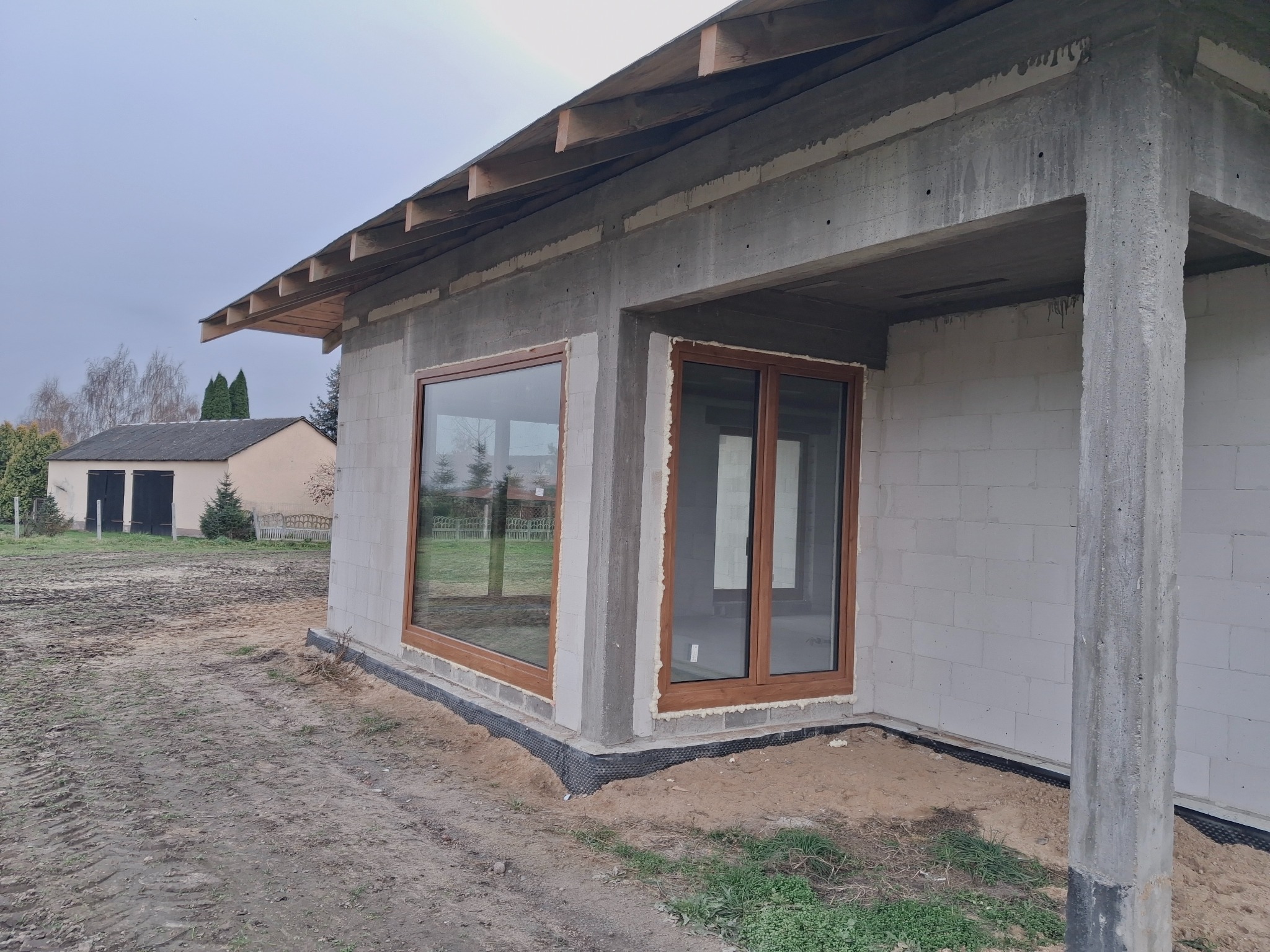 Montaż okien z brązowymi ramami w surowym budynku z betonu komórkowego. Widoczne detale montażu i otoczenie placu budowy. Ujęcie z zewnątrz, w pochmurny dzień.