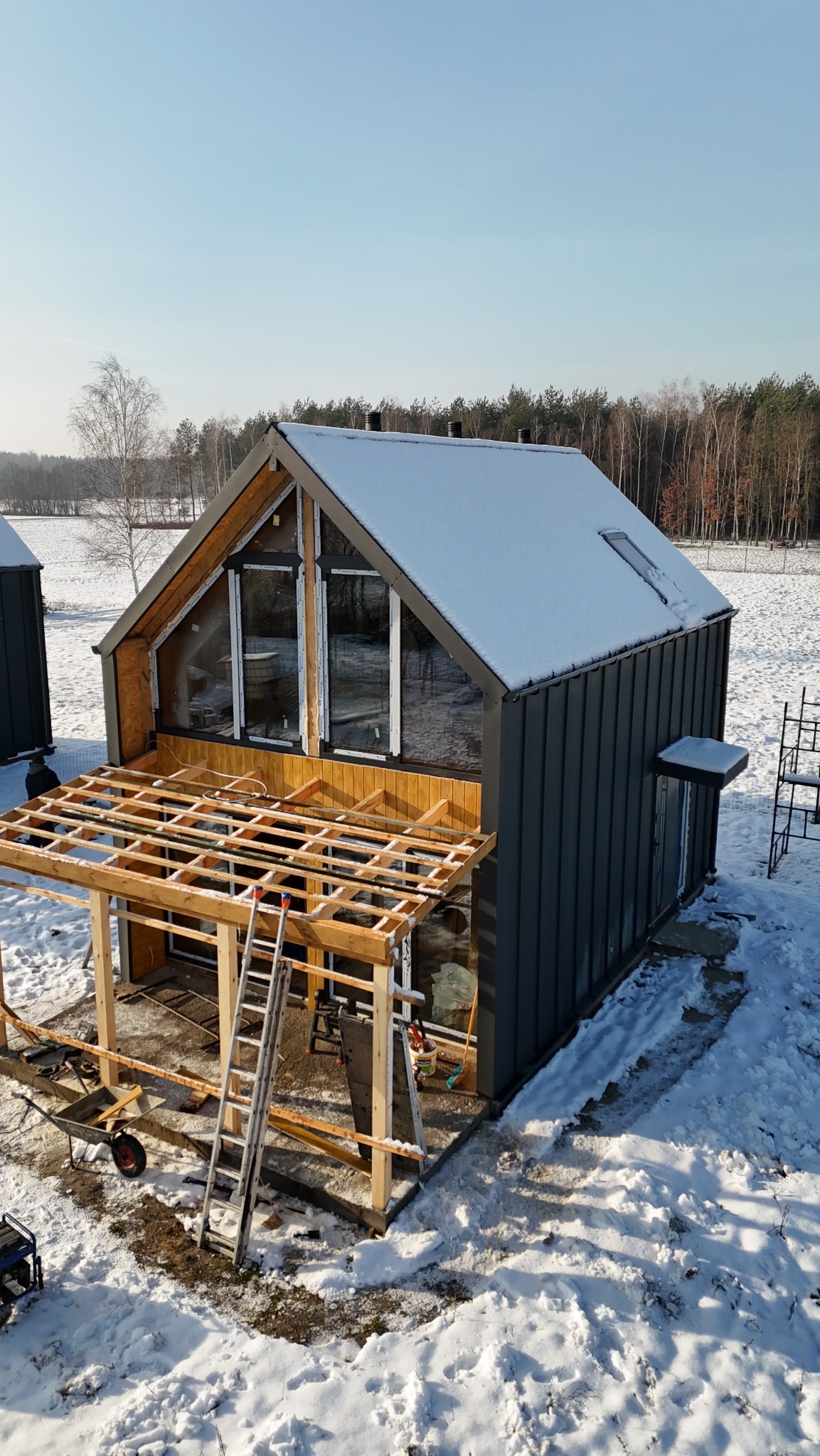 Montaż stolarki okiennej w nowoczesnym domu z szarej blachy na tle zimowego krajobrazu. Widoczne rusztowanie i drabina.