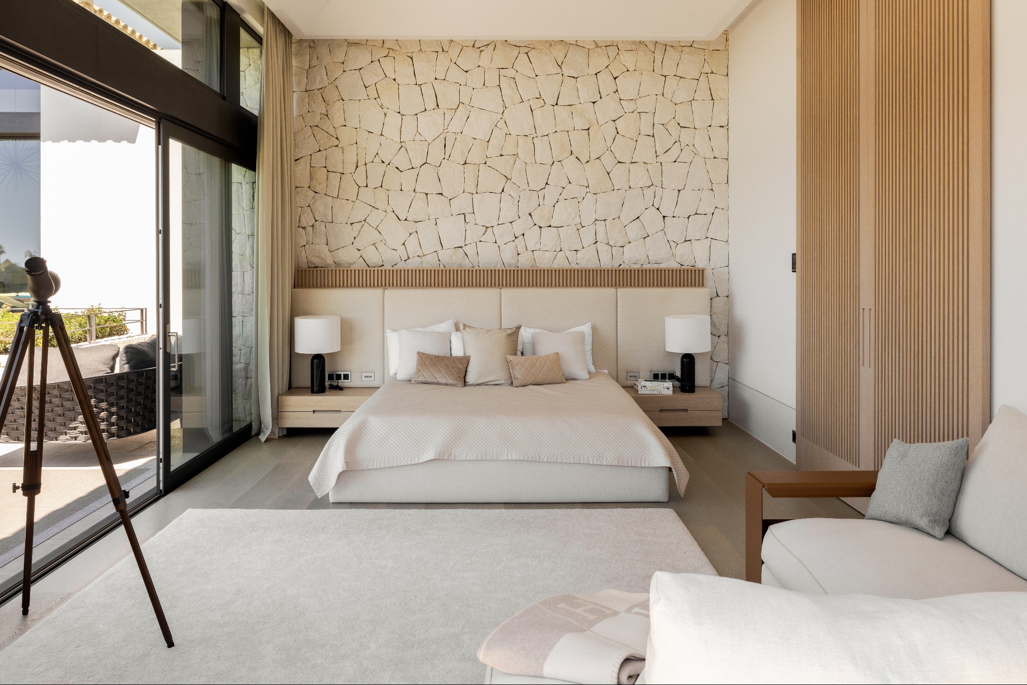 Jasna sypialnia w rezydencji: kamienna ściana za łóżkiem, drewniana szafa, teleskop przy oknie z widokiem na zewnątrz. Beżowa kolorystyka, nowoczesny design.