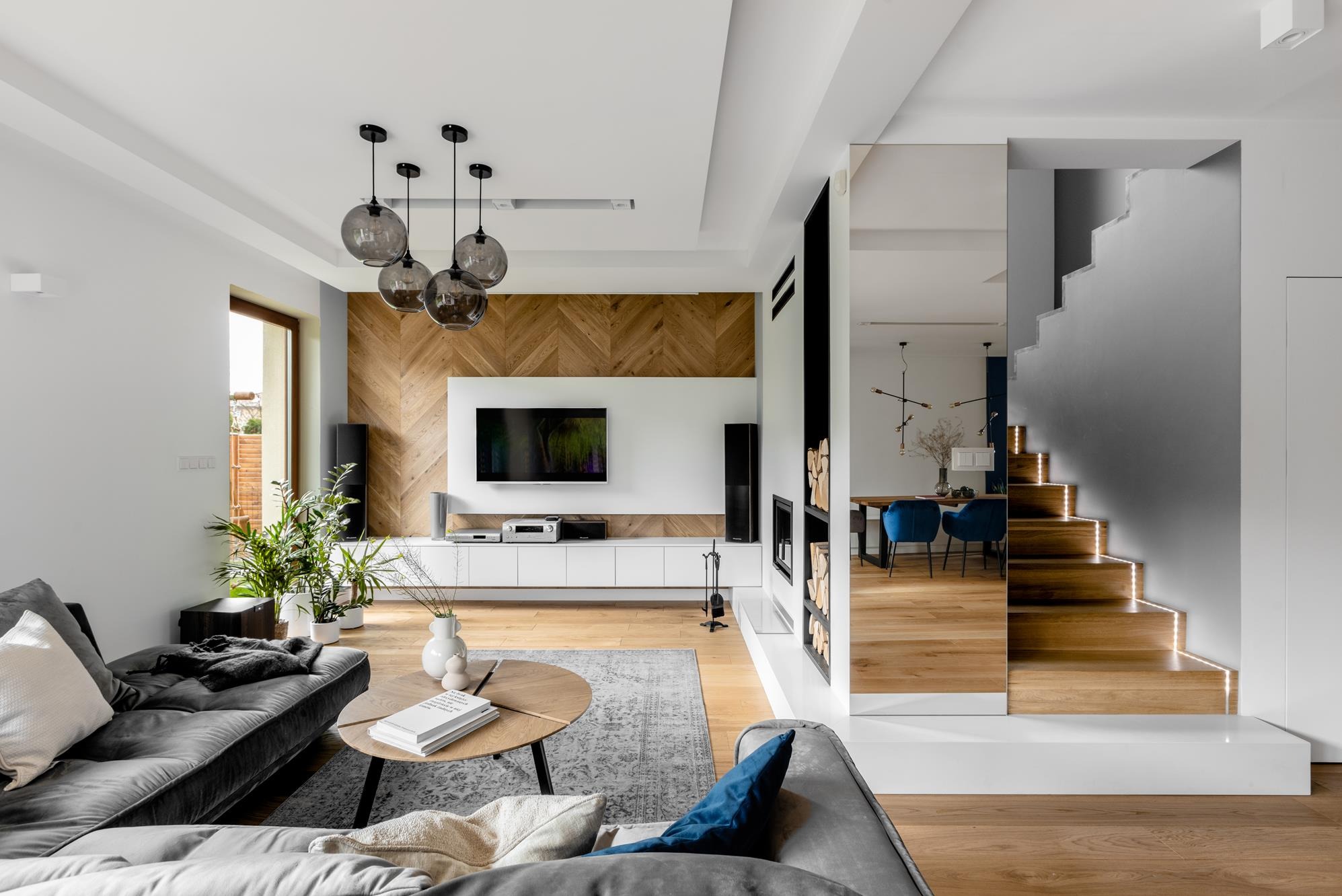 Nowoczesny salon z drewnianą ścianą w jodełkę, szarą sofą i schodami z podświetleniem LED. Lustro powiększa przestrzeń, odbijając jadalnię z niebieskimi krzesłami.