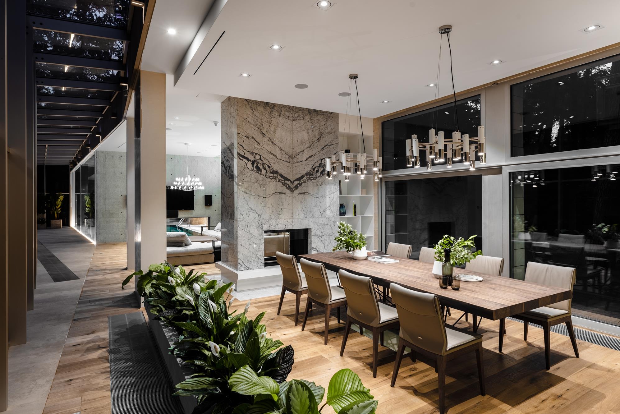 Nowoczesna jadalnia z długim drewnianym stołem, marmurowym kominkiem i dużymi oknami. Rośliny doniczkowe dodają wnętrzu świeżości. Widok na salon z basenem.