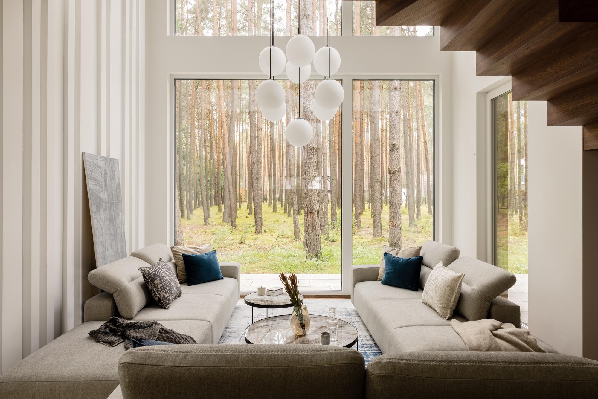 Jasny salon z widokiem na las przez duże okno. Dwie sofy, stoliki kawowe i nowoczesna lampa kulista. Minimalistyczny design z drewnianymi schodami w tle.