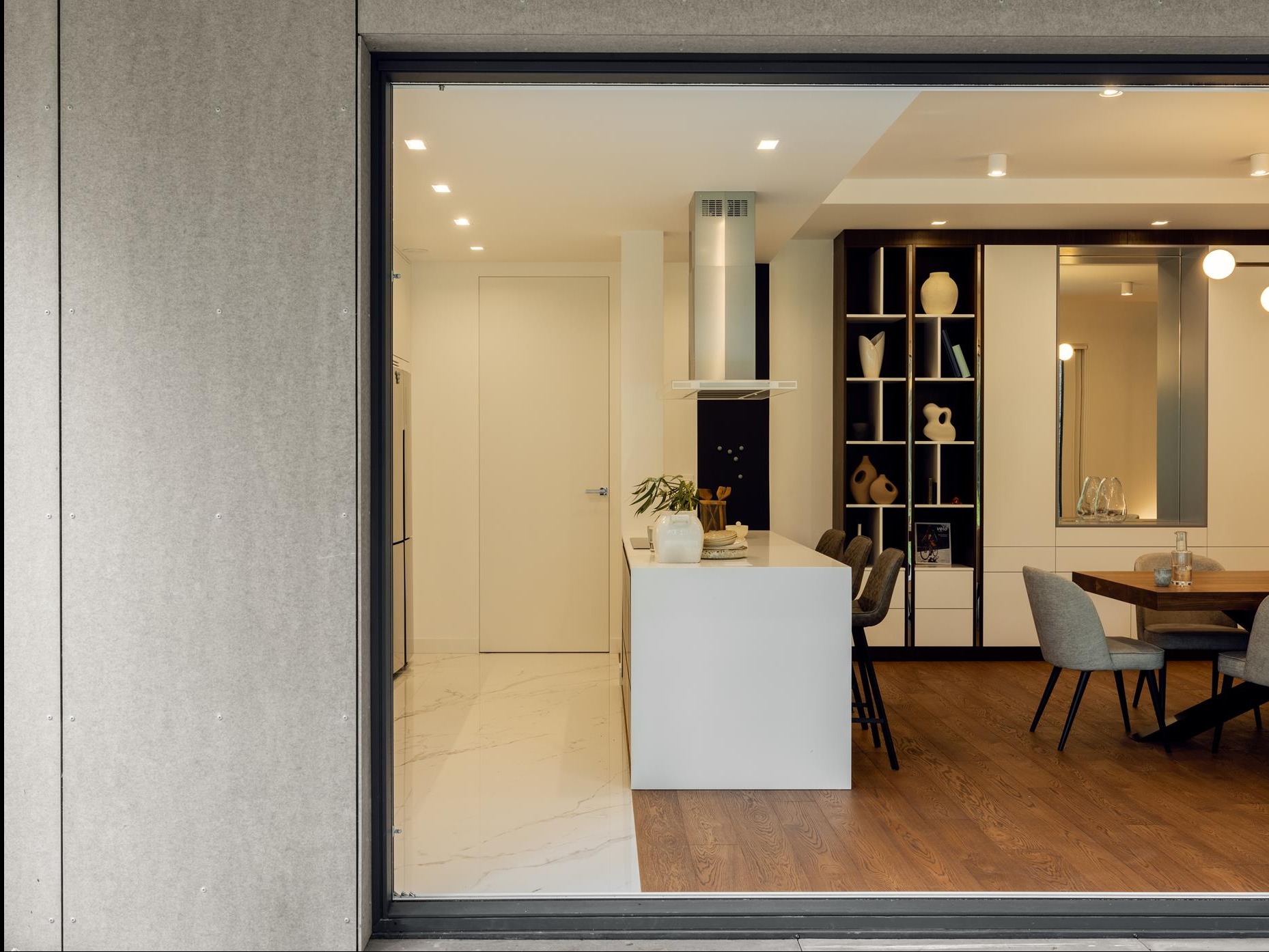 Nowoczesne wnętrze domu z wyspą kuchenną, widoczne przez duże okno. Białe ściany, marmurowa podłoga w kuchni, drewniana w jadalni. Minimalistyczny design, ciepłe oświetlenie.