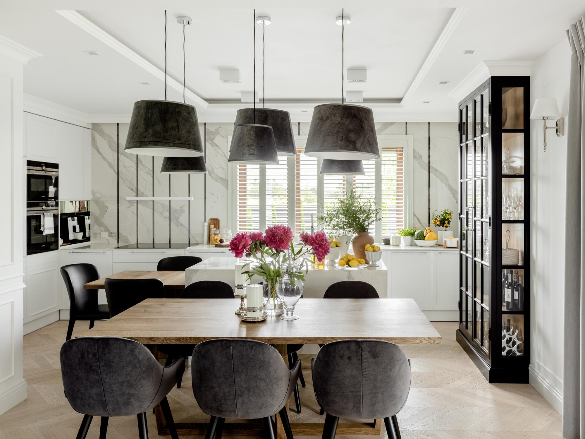 Jasna jadalnia z drewnianym stołem, szarymi krzesłami i marmurową ścianą. Nad stołem wiszą cztery duże, szare lampy. W tle widoczna kuchnia i przeszklona witryna.