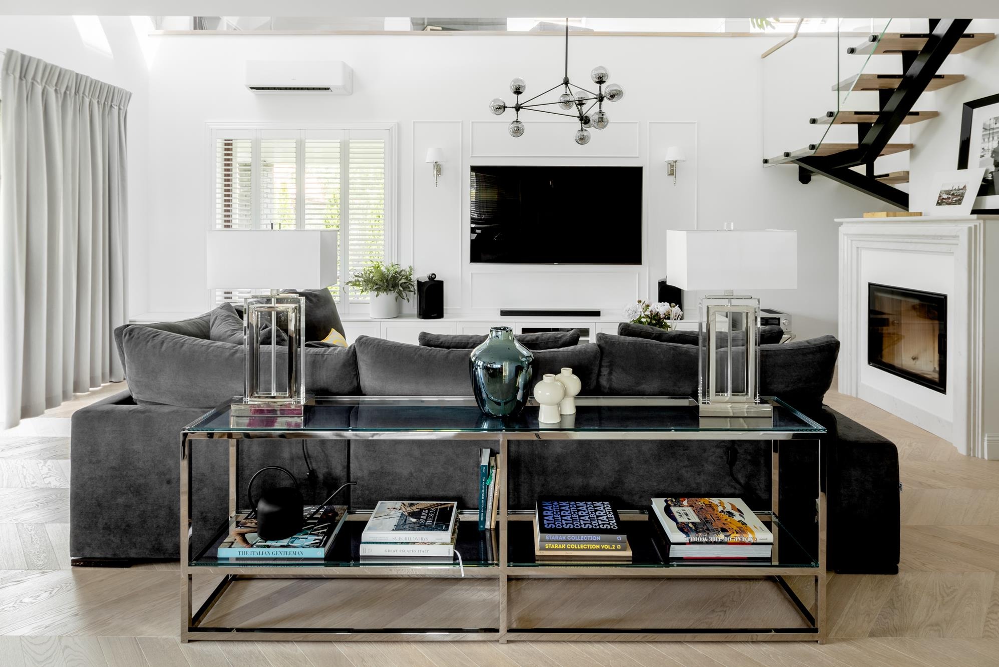 Jasne wnętrze salonu z szarą sofą, szklanym stolikiem kawowym i schodami w tle. Minimalistyczny design z elementami dekoracyjnymi i oświetleniem.