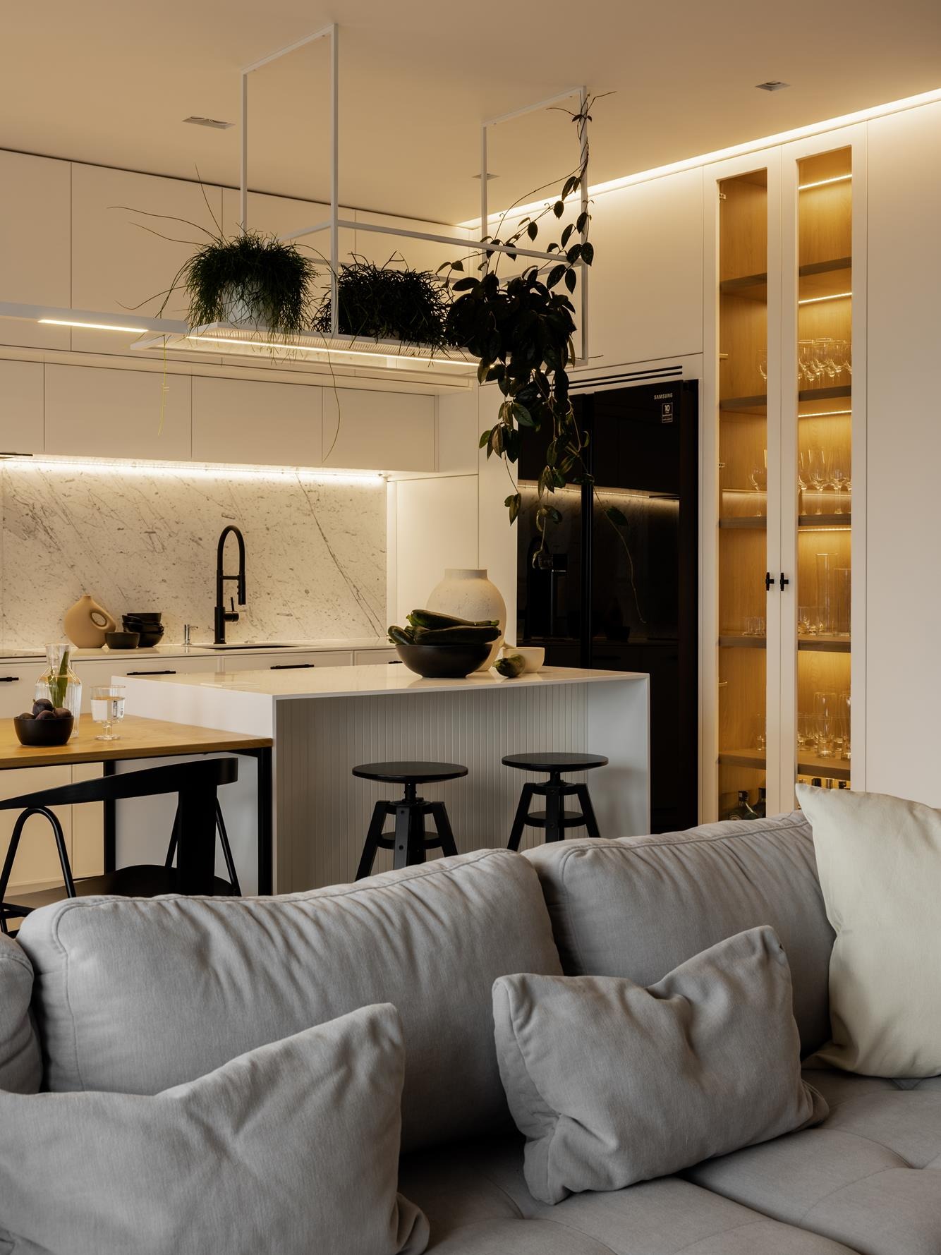 Nowoczesna kuchnia z wyspą i marmurowym blatem, oświetlona LED, z wiszącymi roślinami i szklaną witryną. W tle czarna lodówka Samsung i szara sofa.