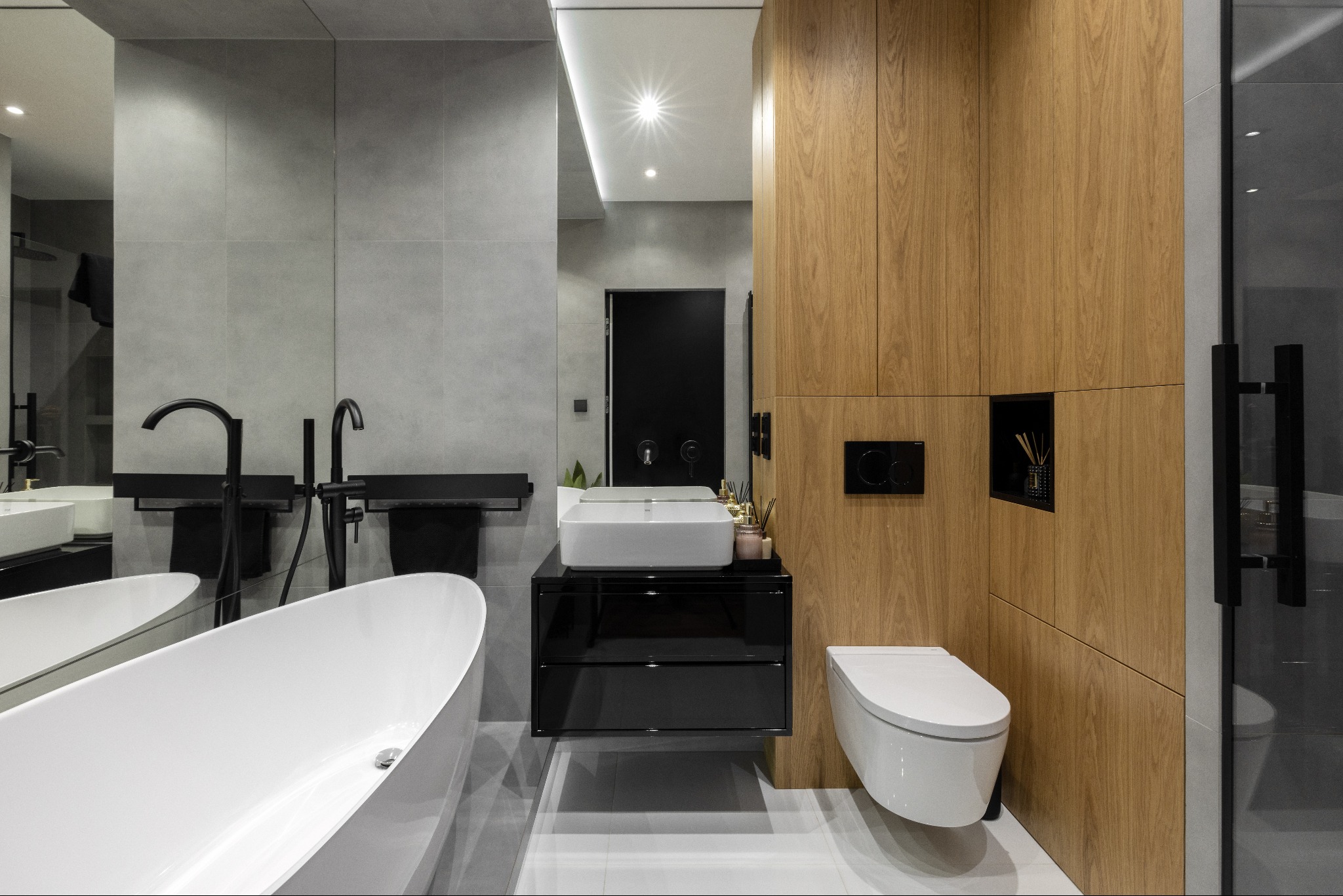 Nowoczesna łazienka z wolnostojącą wanną, umywalką nablatową na czarnej szafce i drewnianą zabudową z toaletą. Szare płytki i czarne akcenty.