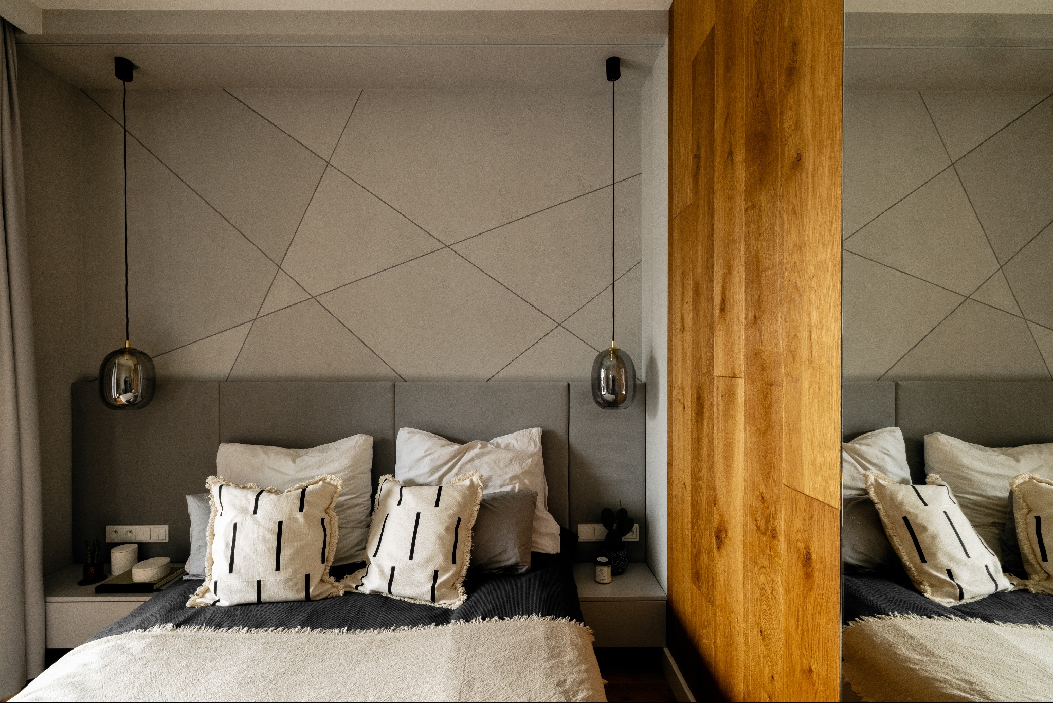 Nowoczesna sypialnia w odcieniach szarości z drewnianą zabudową. Geometryczny wzór na ścianie, wiszące lampy i dekoracyjne poduszki tworzą przytulną atmosferę.