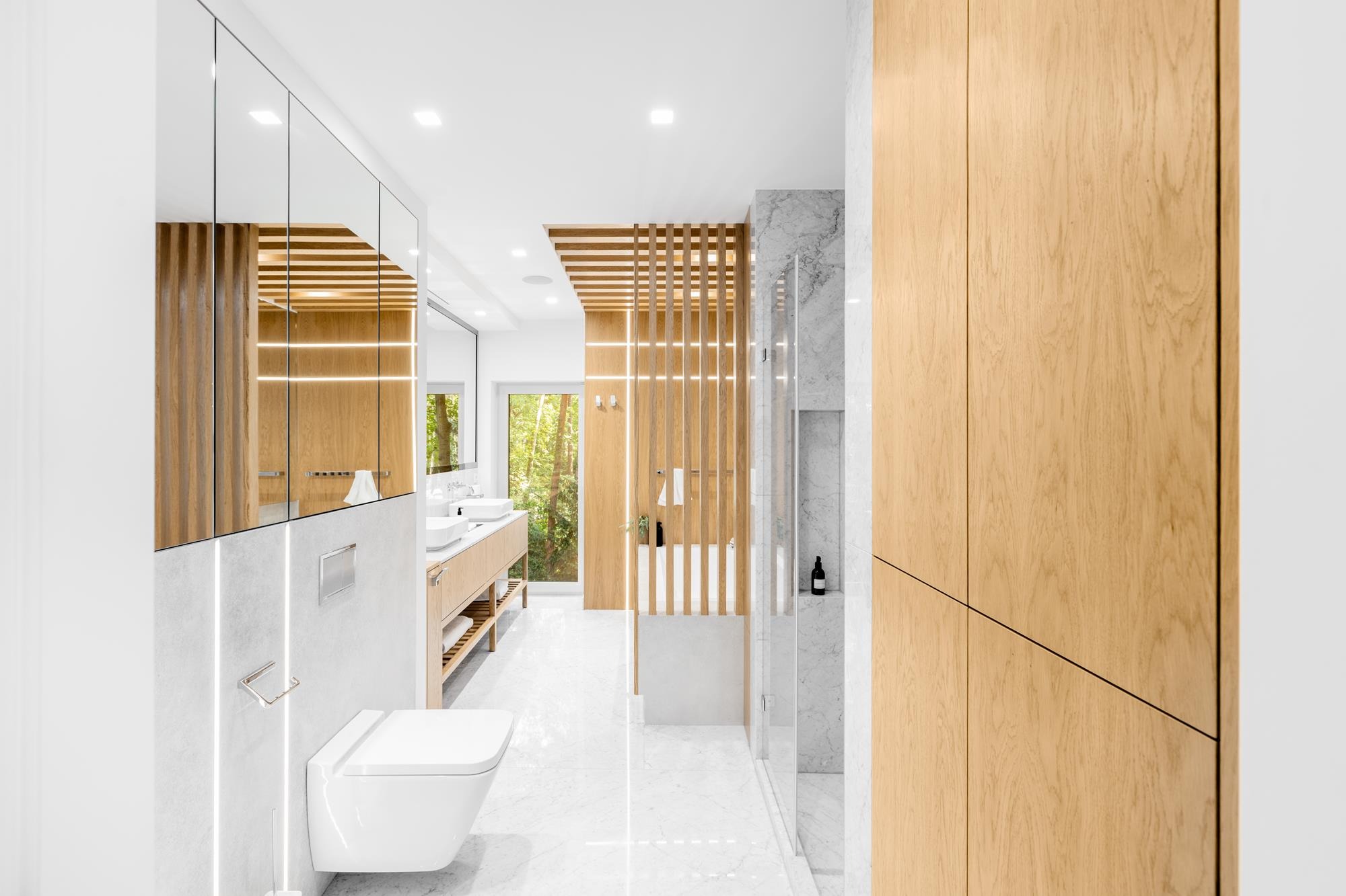 Nowoczesna łazienka z drewnianymi akcentami: szafki z lustrami, podwieszana toaleta, marmurowe płytki i drewniana ścianka działowa z oświetleniem LED.