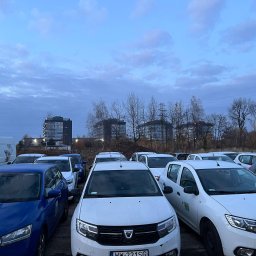 Cabvibe - Flota samochodów Dacia Sandero i Skoda Fabia na parkingu, gotowych do wynajmu dla kierowców, z budynkami w tle i pochmurnym niebem.