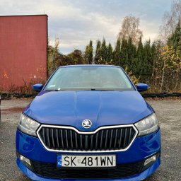 Cabvibe - Niebieska Skoda Fabia z przodu, tablica rejestracyjna SK 481WN, na tle czerwonego budynku i drzew. Auto gotowe do świadczenia usług transportowych.