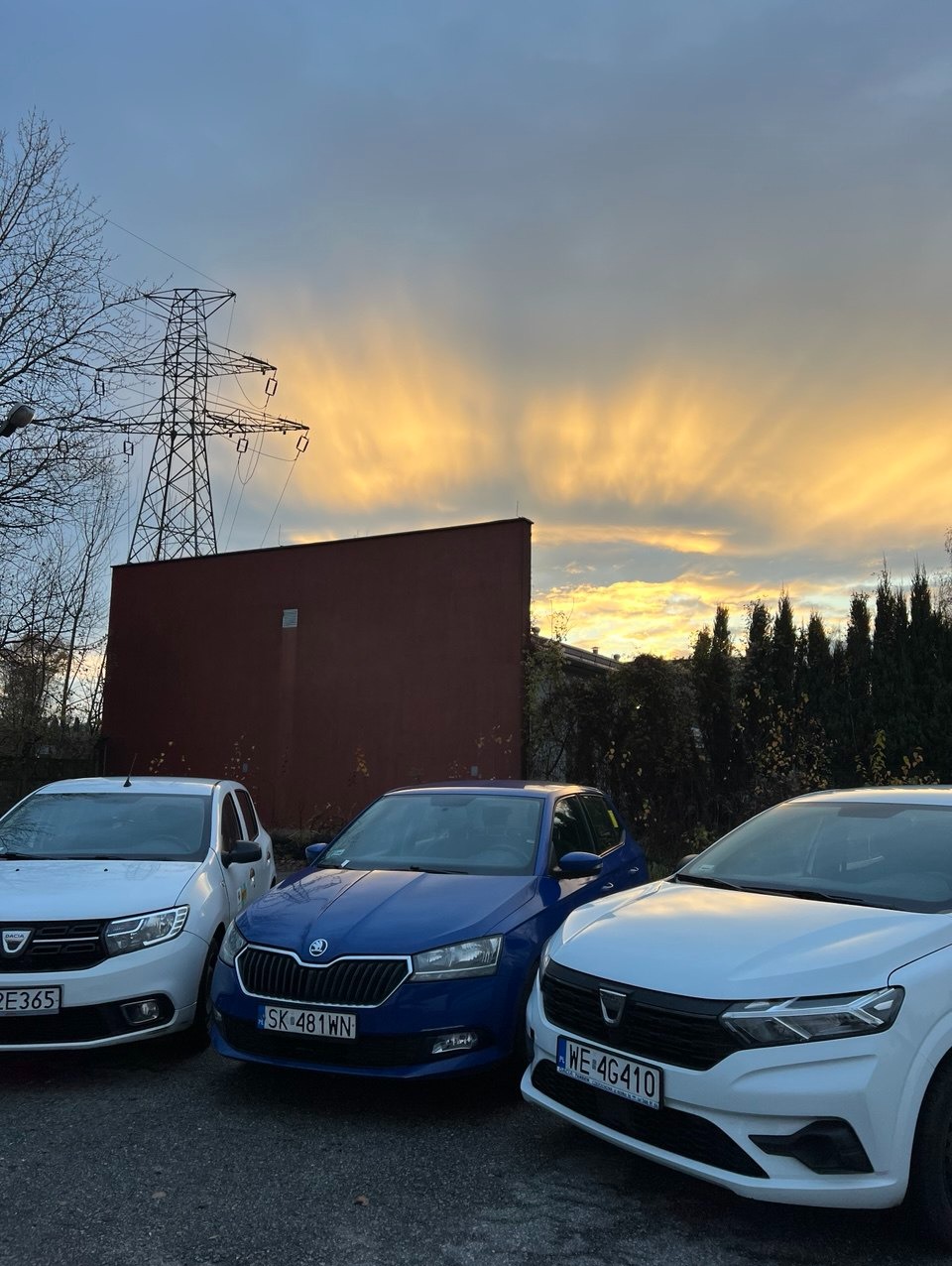 Trzy auta (Dacia, Skoda) na parkingu o zmierzchu. W tle słup wysokiego napięcia i budynek. Dwa białe auta po bokach, niebieskie pośrodku. Widoczne tablice rejestracyjne.