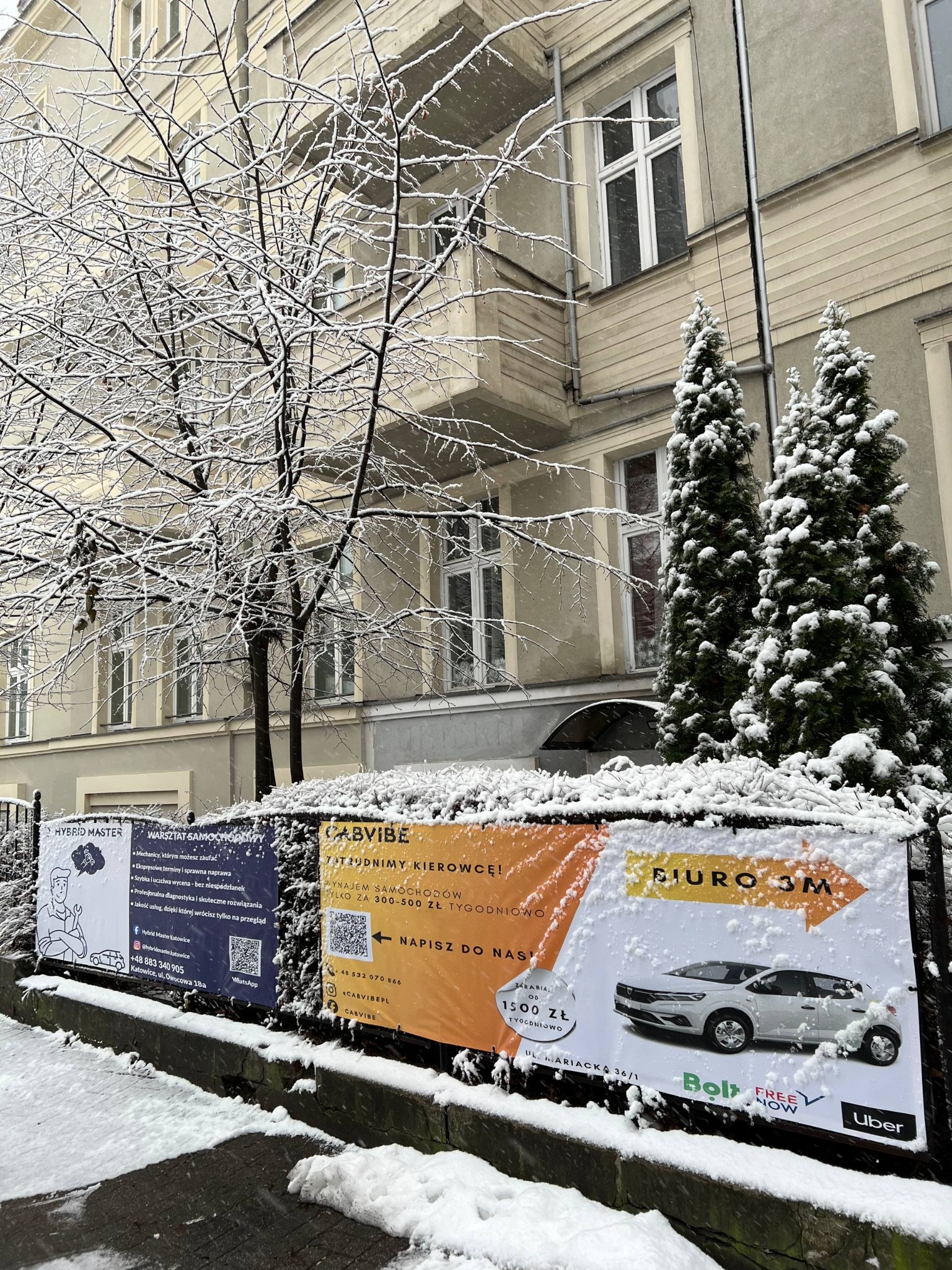Zimowy kadr z reklamami Cabvibe Taxi i Hybrid Master na tle budynku. Śnieg pokrywa plakaty, krzewy i drzewa. Widoczne logo Bolt i Uber.