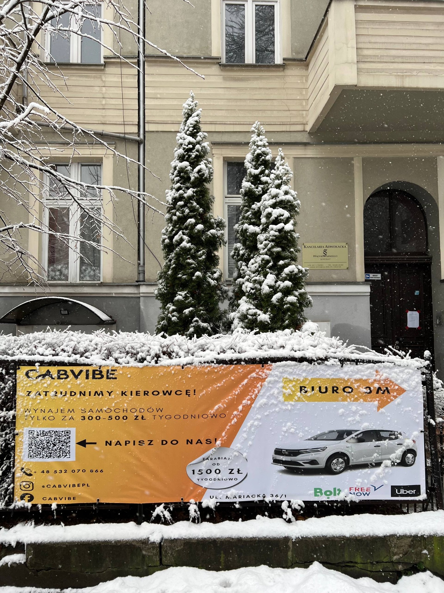 Zimowy plakat CabVibe z ofertą wynajmu samochodów dla kierowców, zlokalizowany przed budynkiem w Katowicach. Śnieg na plakacie i otoczeniu.