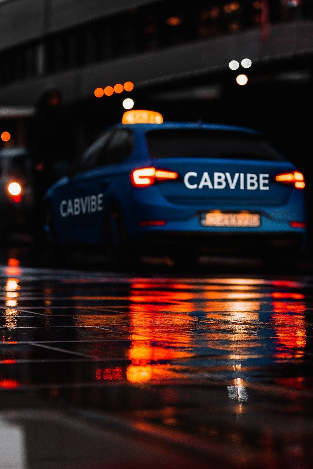 Błękitny samochód CabVibe z włączonymi światłami stopu, widziany z tyłu na mokrej nawierzchni odbijającej pomarańczowe światła miasta nocą.
