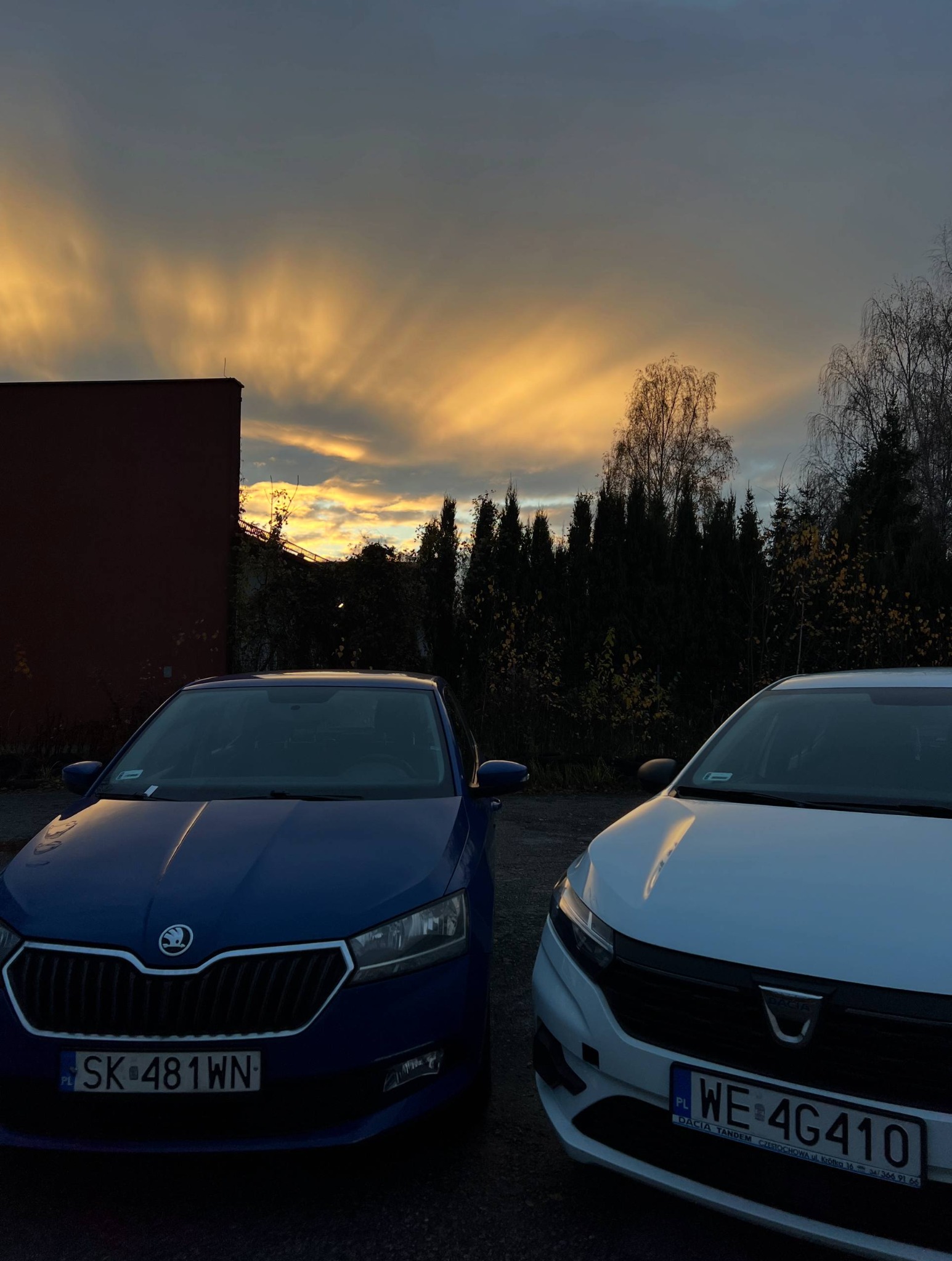 Dwa samochody: niebieska Skoda i biała Dacia, zaparkowane na tle budynku i drzew, z dramatycznym, zachodzącym słońcem na niebie. Auta gotowe do pracy w aplikacjach taxi.