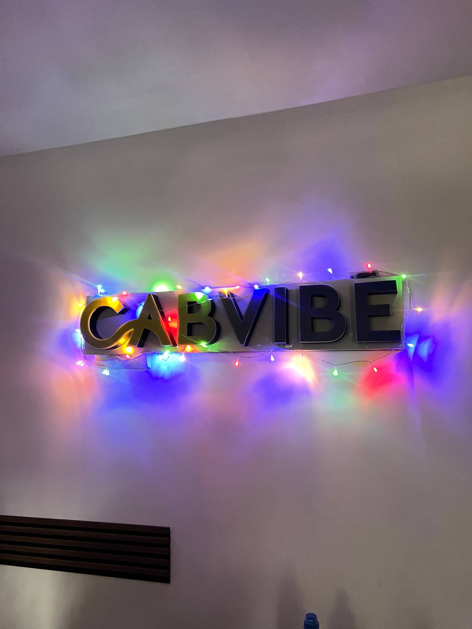 Podświetlany napis 'CABVIBE' na ścianie, otoczony kolorowymi lampkami choinkowymi. Litery w kolorach złotym i ciemnoszarym. Dekoracja wnętrza.