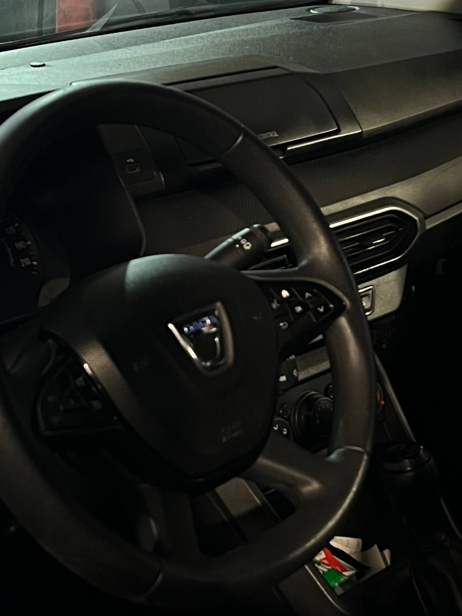 Wnętrze samochodu Dacia Sandero 2021 z widoczną kierownicą, deską rozdzielczą i elementami sterowania. Ujęcie z perspektywy kierowcy, ciemne oświetlenie.