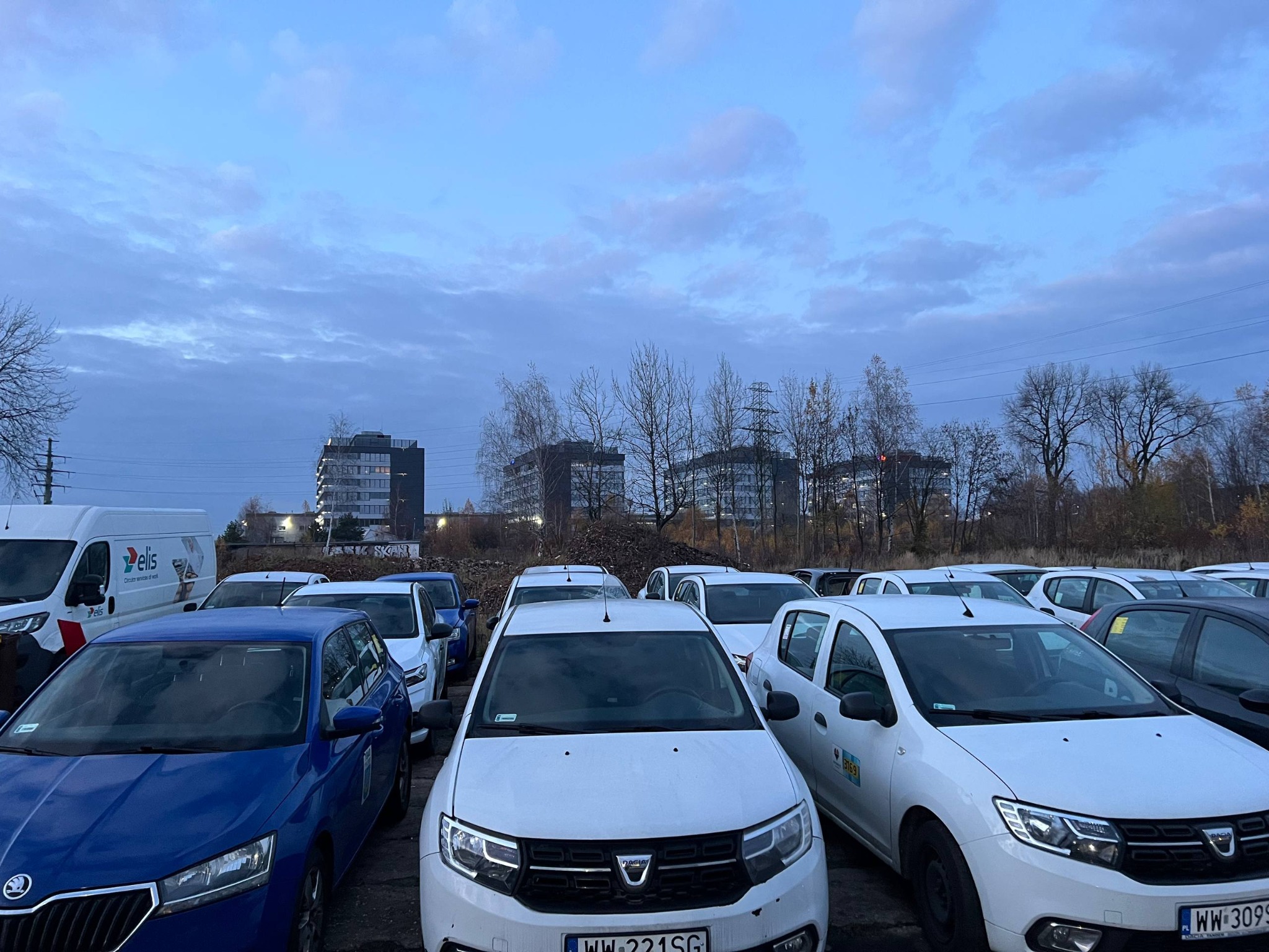 Flota samochodów Dacia Sandero i Skoda Fabia na parkingu, gotowych do wynajmu dla kierowców, z budynkami w tle i pochmurnym niebem.