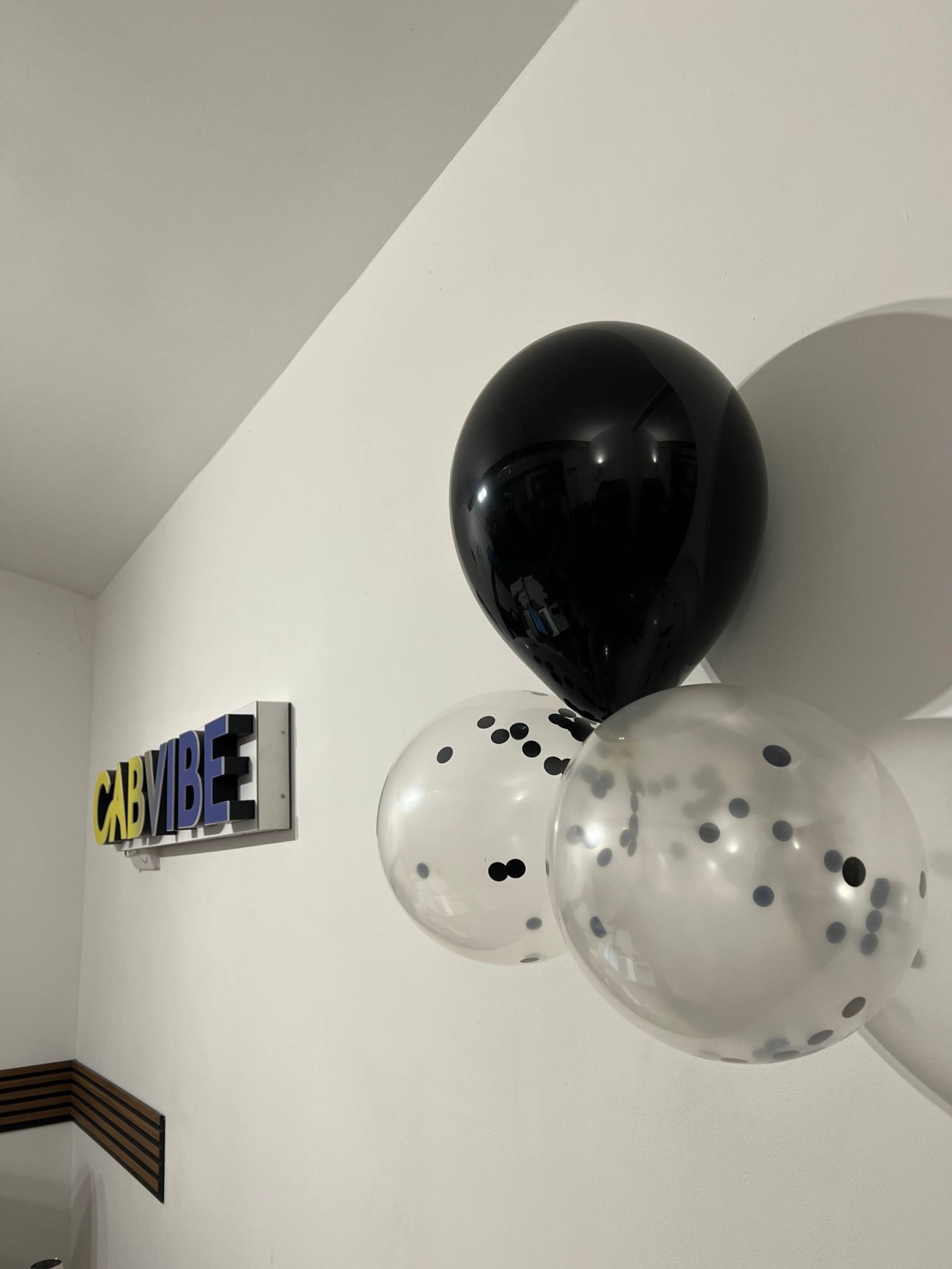 Trzy balony (czarny i dwa przezroczyste z konfetti) wiszą na białej ścianie obok podświetlanego logo firmy taksówkarskiej 'CABVIBEE'.