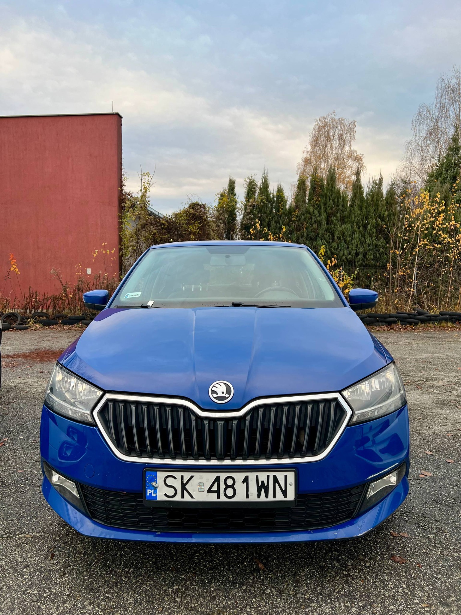 Niebieska Skoda Fabia z przodu, tablica rejestracyjna SK 481WN, na tle czerwonego budynku i drzew. Auto gotowe do świadczenia usług transportowych.