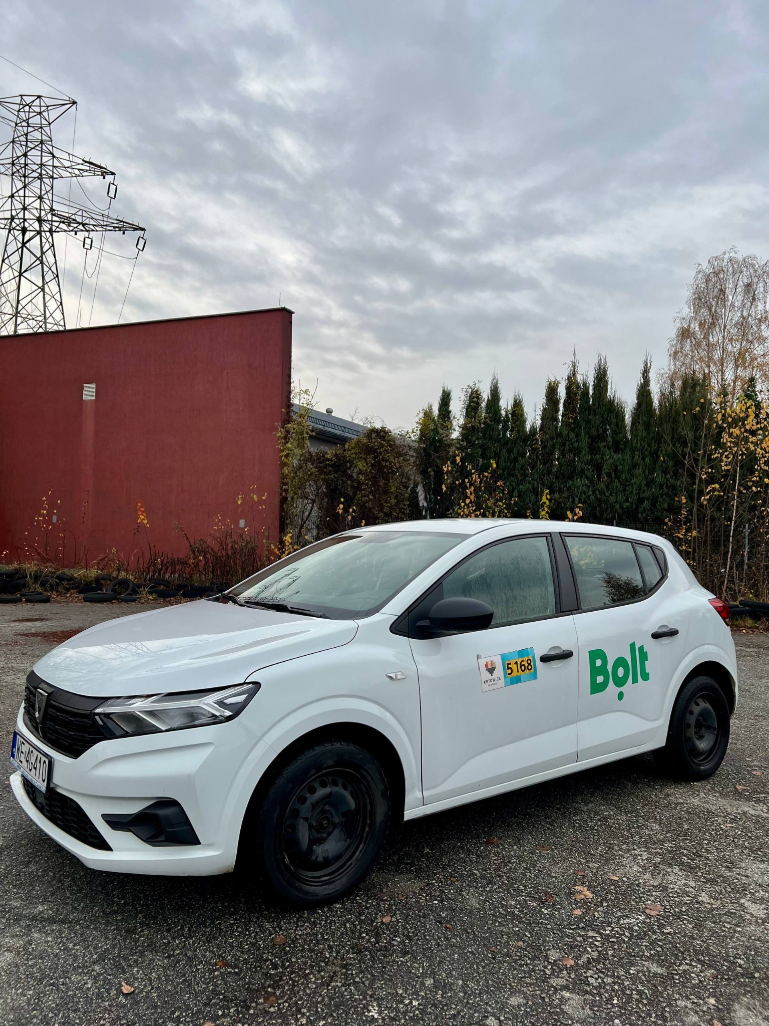 Biała Dacia Sandero z logo Bolt na parkingu, widok z boku. W tle słup wysokiego napięcia i czerwony budynek. Auto z czarnymi felgami i numerem bocznym.