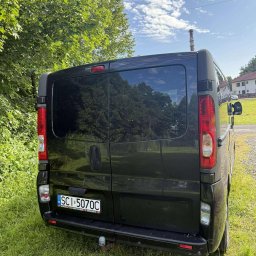 FHU KLAUDIA KŁODA - Czarny Opel Vivaro z tyłu, zaparkowany na trawie. Widoczny hak holowniczy i tablica rejestracyjna. W tle drzewa i budynek.