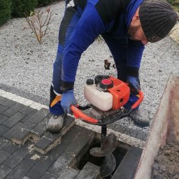 FHU KLAUDIA KŁODA - Pracownik w kombinezonie obsługuje wiertnicę spalinową, wiercąc otwór w kostce brukowej. Obok taczka z ziemią. W tle żywopłot i ogrodzenie.
