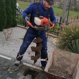 FHU KLAUDIA KŁODA - Mężczyzna wierci otwór w ziemi wiertnicą spalinową na kostce brukowej. Obok taczka z ziemią. W tle ogrodzenie i zieleń. Praca w ogrodzie.