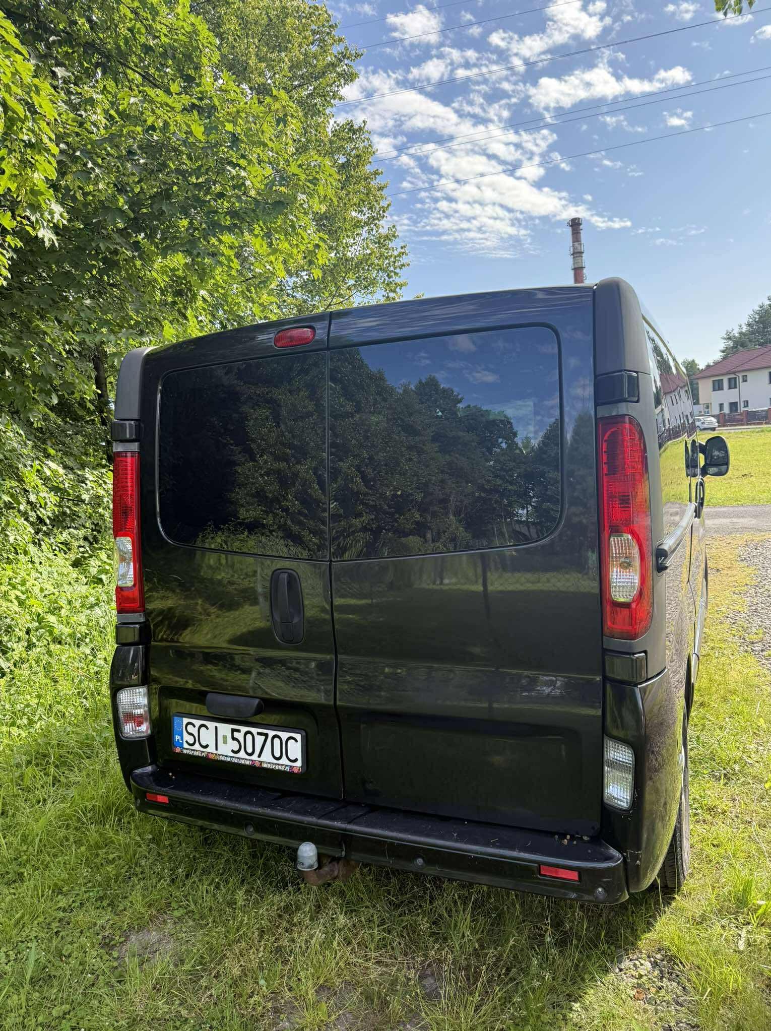 Czarny Opel Vivaro z tyłu, zaparkowany na trawie. Widoczny hak holowniczy i tablica rejestracyjna. W tle drzewa i budynek.