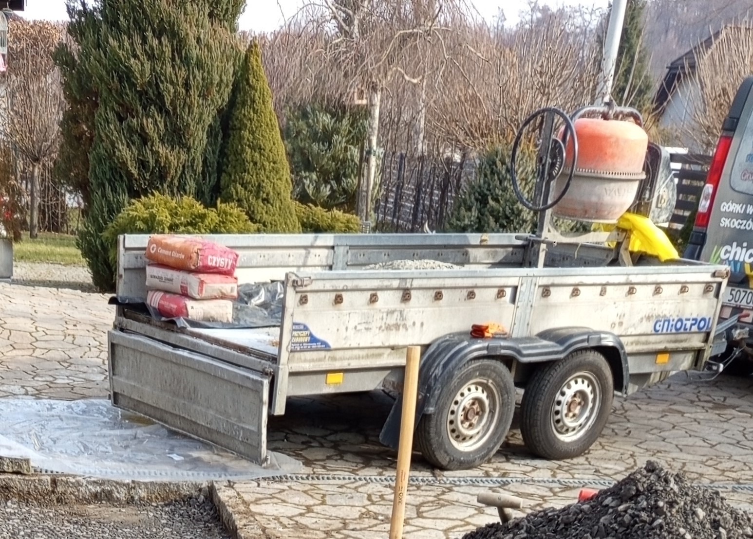 Przyczepa GNIOTPOL z betoniarką i workami cementu na placu budowy. Widoczny fragment chodnika z kostki i sterta gruzu. Dzień.
