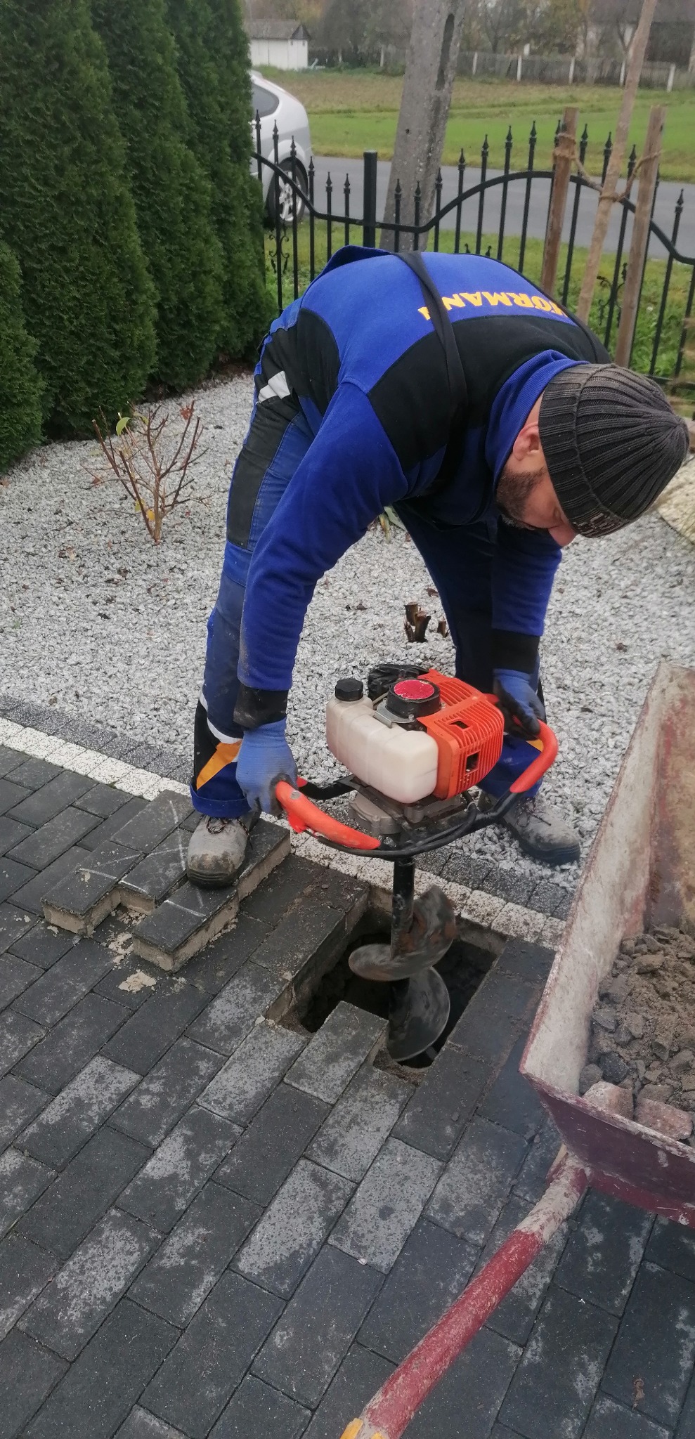 Pracownik w kombinezonie obsługuje wiertnicę spalinową, wiercąc otwór w kostce brukowej. Obok taczka z ziemią. W tle żywopłot i ogrodzenie.